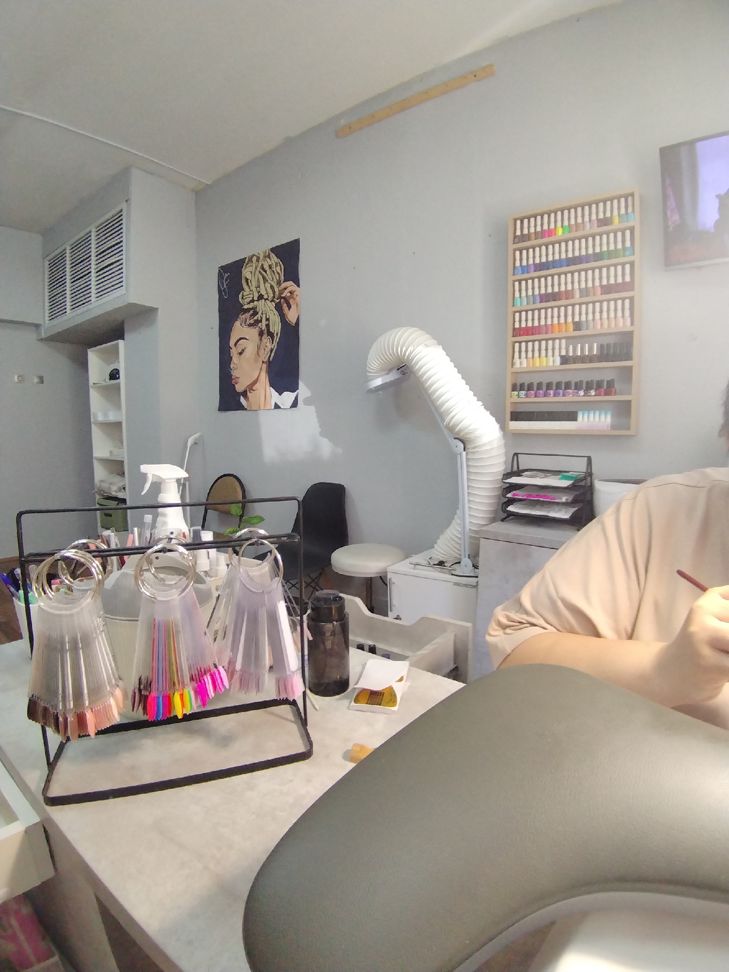 The image depicts the interior of a nail salon. In the foreground, there's a manicure station with a gray armrest and a stand holding nail polish color swatches. A spray bottle and other salon supplies are also visible on the station. In the background, there's a poster of a woman with braided hair, a shelf displaying nail polish bottles, and a white ventilation pipe. The walls are painted gray, and there is a person to the right of the image partially visible. The environment appears to be a typical commercial space for cosmetic services. This analysis lacks information needed to specifically identify the location in Noviy, Russia.