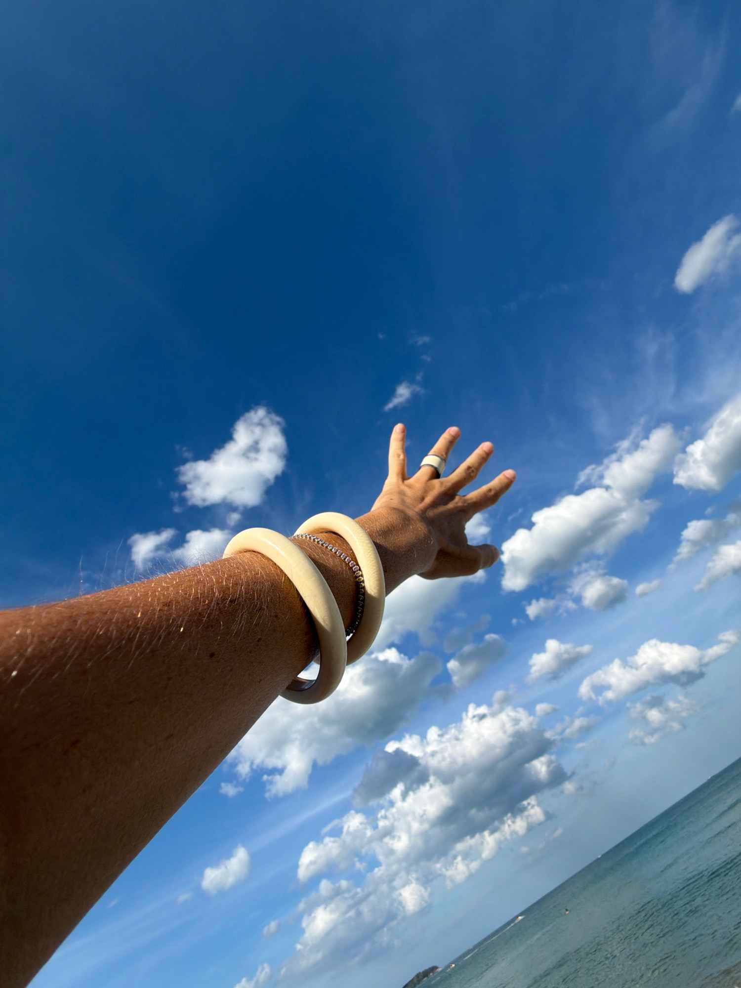 In the city of Ko Samui District, Thailand, a person's arm and hand extend upwards towards a vibrant blue sky dotted with fluffy white clouds. The arm is adorned with two large, cream-colored bangles and a thin, sparkly bracelet. A ring is visible on the index finger of the outstretched hand, which is splayed open with fingers extended. Below the arm and hand, a turquoise ocean meets the horizon, with a faint indication of land on the far left. The perspective of the shot is from below, looking up at the sky and the person's limb.