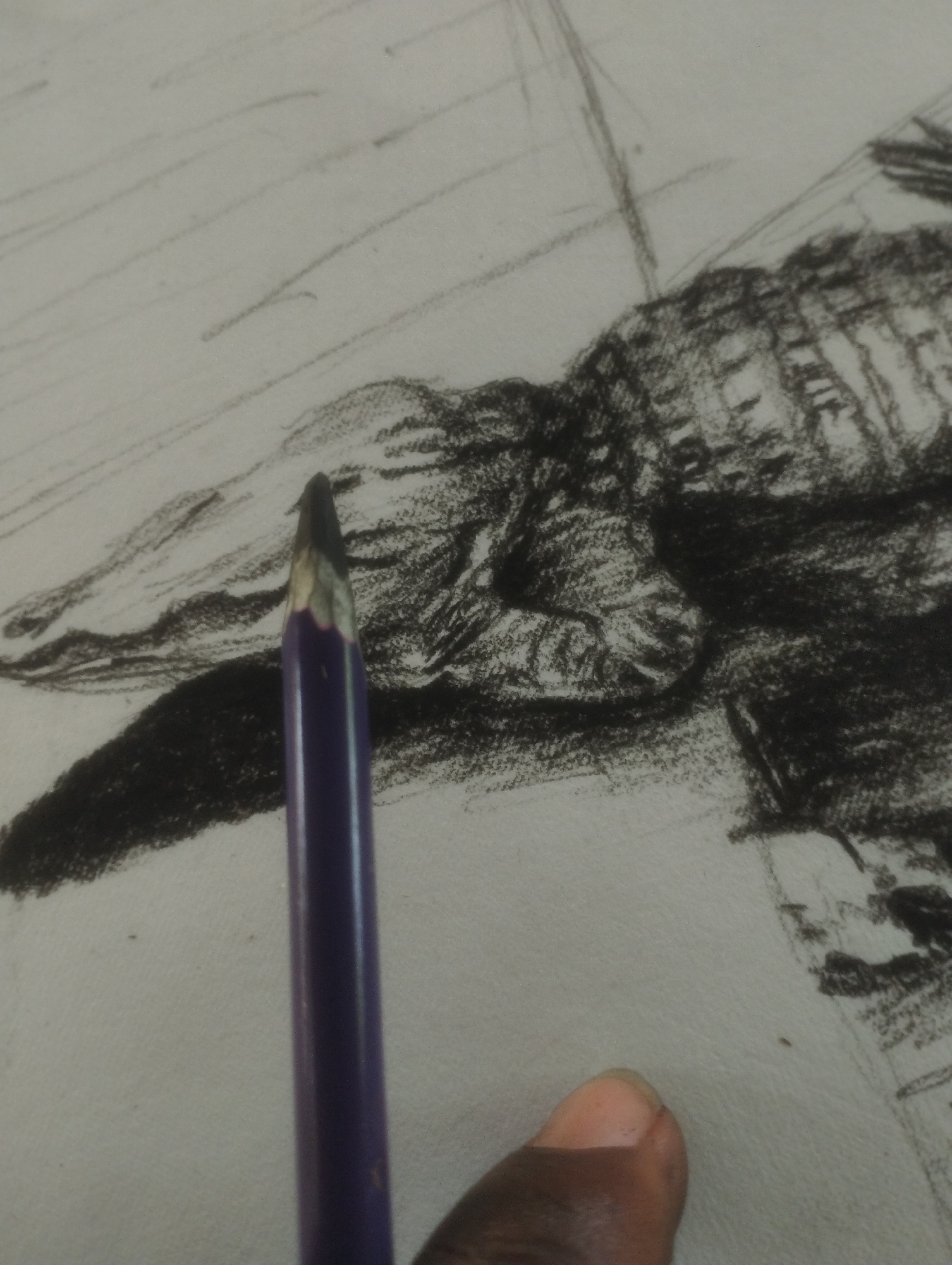 The image shows a close-up view of a charcoal sketch on paper. The sketch appears to be of a figure, possibly a person, rendered in dark, smudged tones with some lighter lines indicating form. In the foreground, a purple pencil with a sharpened graphite tip is positioned vertically, its point just above the paper. A person's thumb is visible in the bottom right corner, appearing to be holding the pencil or gesturing towards the sketch. The background of the sketch features a series of diagonal lines, suggesting texture or movement. The overall impression is one of an artistic process, with the sketch in progress and the tools of creation at hand. No specific time of day, weather, location, or text is discernible.