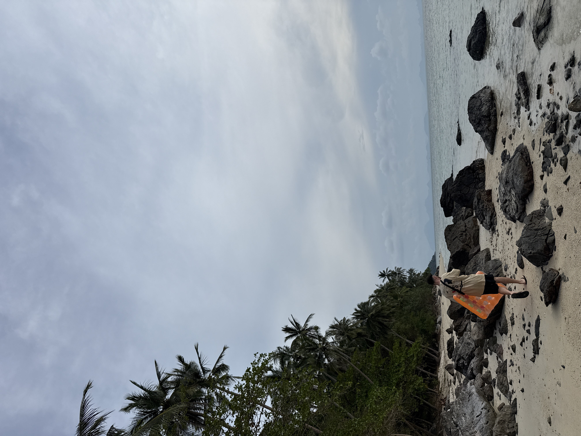 The image depicts a serene tropical beach scene in Baan Pang Ka, Thailand, under an overcast sky. The upper and left portions of the frame are dominated by a soft, light blue-grey sky filled with subtle cloud formations, suggesting either an early morning, late afternoon, or simply a cloudy day.
The horizon line separates the cloudy sky from the calm, grey-toned sea, which extends across the middle and right side of the image. The sea surface shows minimal ripples. Along the water's edge and slightly inland, a sandy beach is visible, heavily interspersed with numerous dark grey and black rocks of varying sizes.
On the left side of the image, inland from the beach, a dense line of lush green tropical foliage is present, featuring many tall palm trees with their distinctive fronds.
A single person, an adult male, is positioned on the rocky sand in the lower right quarter of the image. He is wearing a light-colored short-sleeved top, dark shorts, and dark sandals. He is looking out towards the sea. Slung over his shoulder is a partially deflated or folded bright orange rectangular inflatable float or raft.
The scene conveys a peaceful atmosphere, indicative of a leisurely day at a secluded beach. No discernible text is visible within the image.