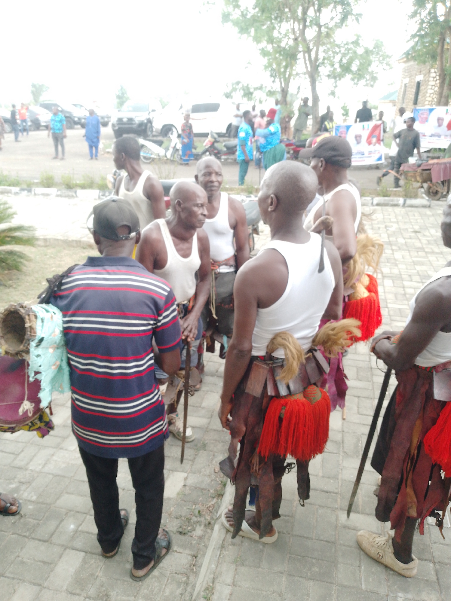 The image captures a gathering of individuals in an urban outdoor setting.
In the foreground, a group of men stands together. Several of the men are wearing white tank tops and distinctive traditional attire, including fringe and leather-like garments around their legs. They appear to be holding staffs or walking sticks. One man in a striped shirt and cap stands slightly apart, facing the group, and is carrying a drum.
In the background, there are more people, some vehicles, and a building with banners or signage. The overall scene suggests a cultural event or gathering in a public space.