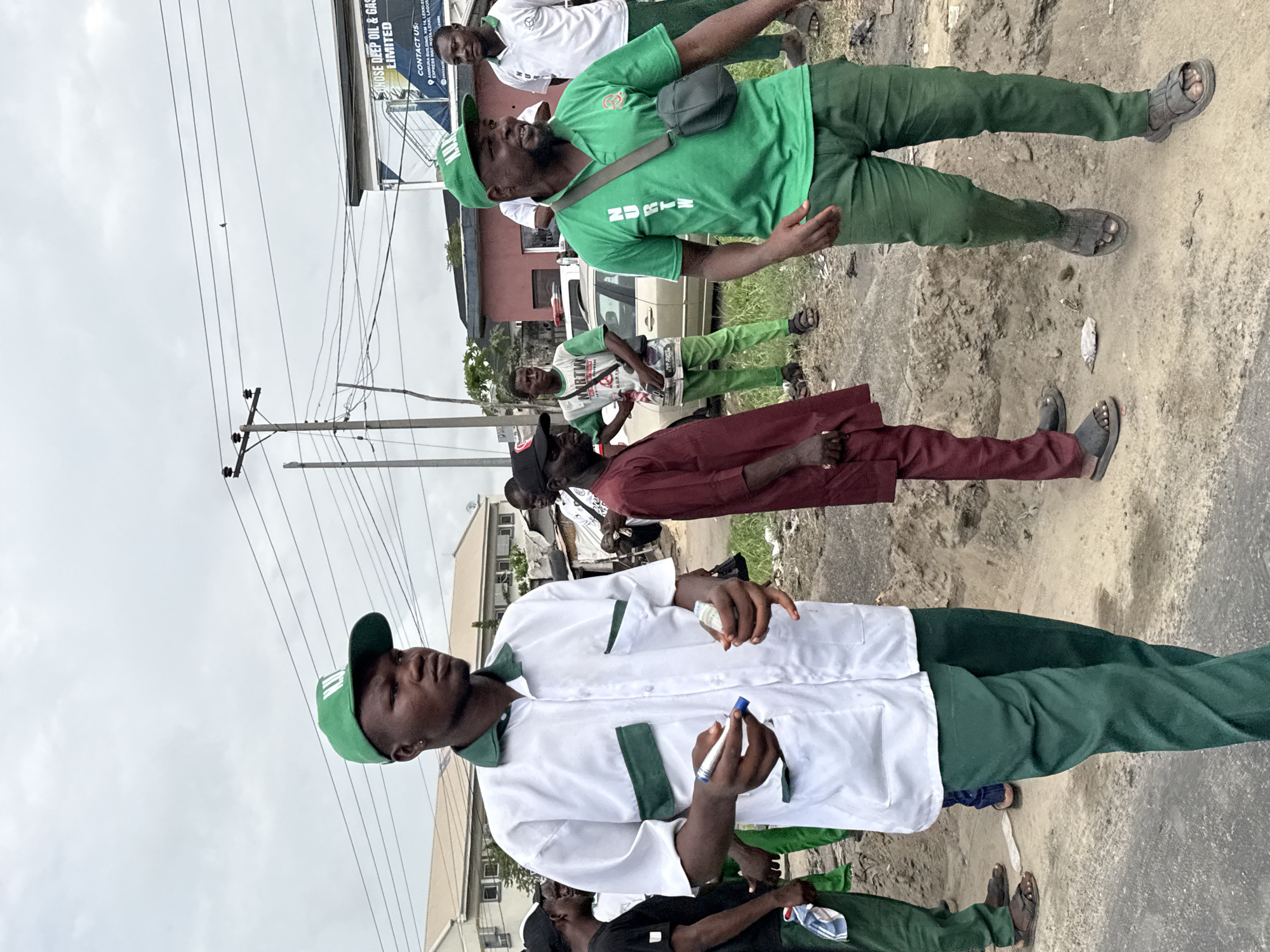 The image depicts a group of people, seemingly men, standing on a paved or dirt road in what appears to be an urban or semi-urban environment. Some individuals are dressed in matching green outfits, including pants, shirts, and caps. One man wears a white shirt with green trim, holding a pen or marker in his hand. A building with a sign reading "ONOSE DEEP OIL & GAS LIMITED, CONTACT US: EXPRE BUILDING, KM 14, LEKKI" suggests the location as Lekki, Nigeria. Other people are dressed in different attire, including a maroon robe-like outfit. The environment includes a cloudy sky, utility poles, and other buildings in the background. Some individuals are wearing sandals.