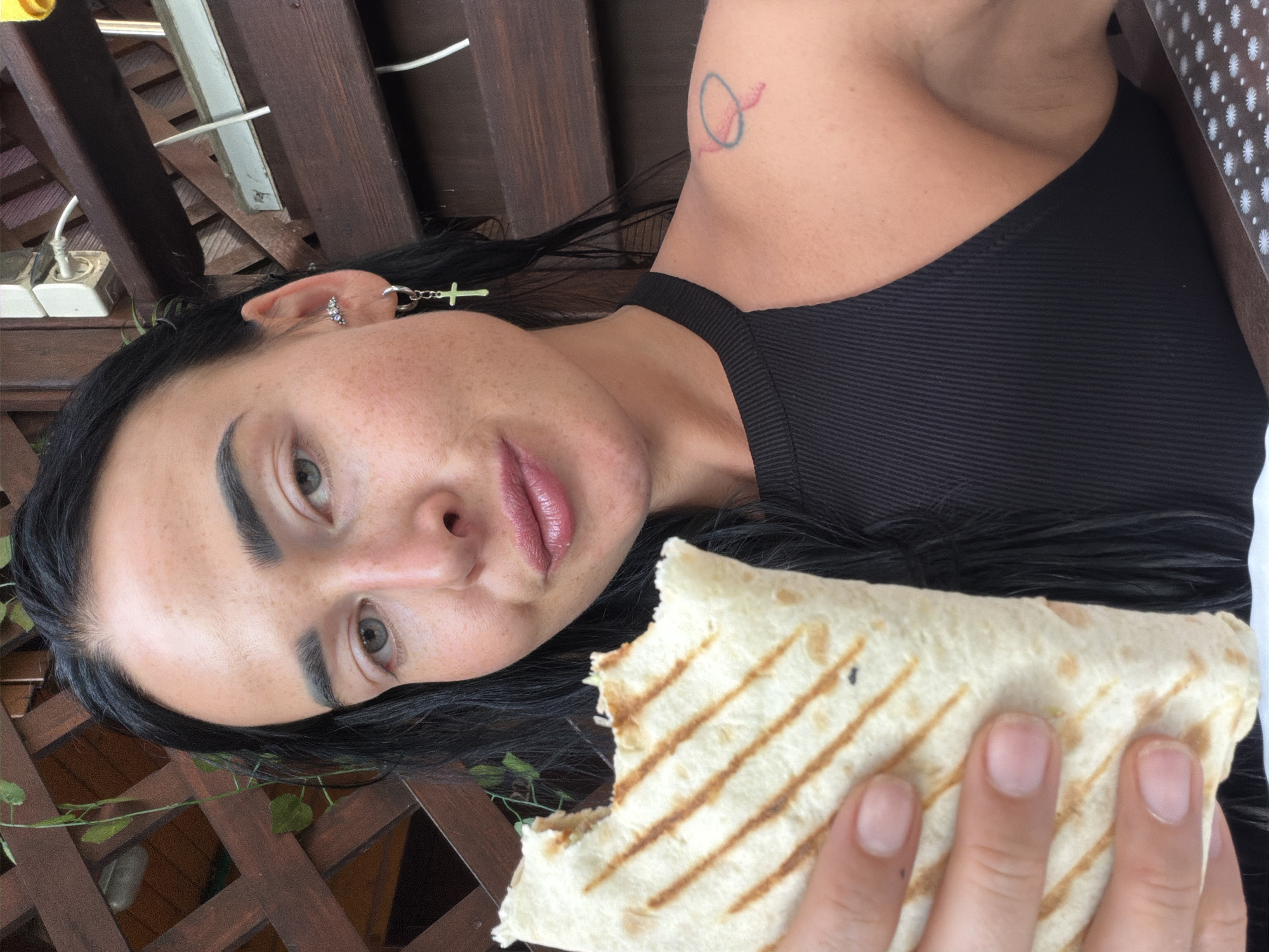 This is a close-up, rotated image of a young woman holding a partially eaten wrap. The woman has long, dark hair, distinctive eyebrows, and light-colored eyes. She is wearing a black ribbed tank top and has several piercings, including a cross earring and smaller stud earrings. There is a small, circular tattoo with red and blue lines on her upper arm. Her skin has a light dusting of freckles.
The wrap appears to be grilled, with visible grill marks on the tortilla. It is held in her right hand, with her fingers visible. The background is blurry but suggests an outdoor setting with wooden slats, possibly a deck or pergola. There is also some greenery visible in the lower left corner. The image appears to be taken during the daytime, likely outdoors. The overall tone is casual. No visible text is present.