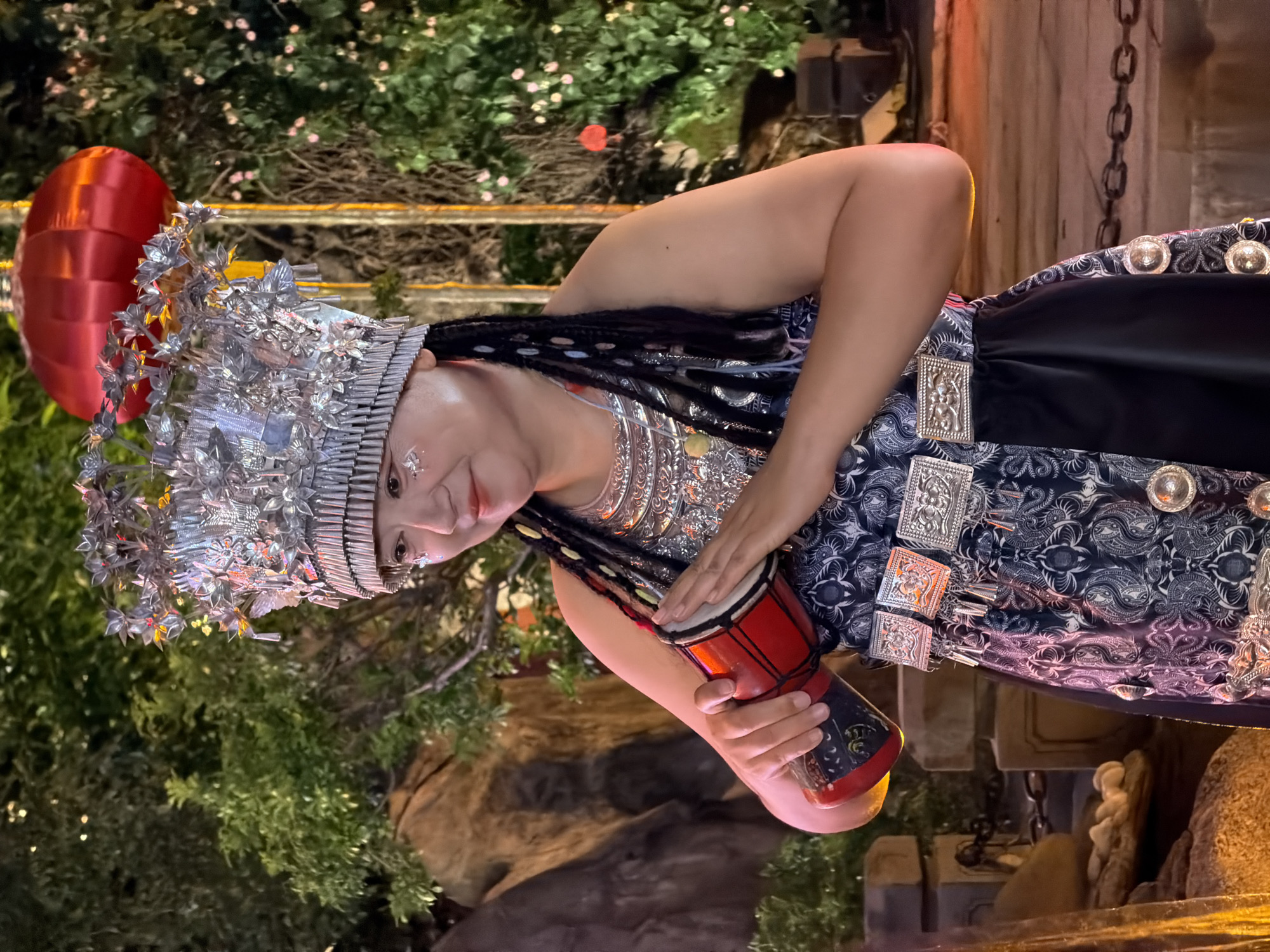 A medium shot captures a woman in traditional attire, holding a red and black miniature drum. She wears an ornate silver headdress adorned with floral and butterfly motifs, and intricate silver necklaces. Her dark hair is braided with colorful accents, and she has decorative glitter on her cheeks. She is dressed in a dark blue patterned top and a black skirt with decorative silver embellishments at the waist and hem. She stands in an outdoor setting, with lush green foliage and decorative lights visible in the background, along with a large red lantern. The overall scene appears to be a festive cultural performance or celebration, potentially in Yuzhong District, China.