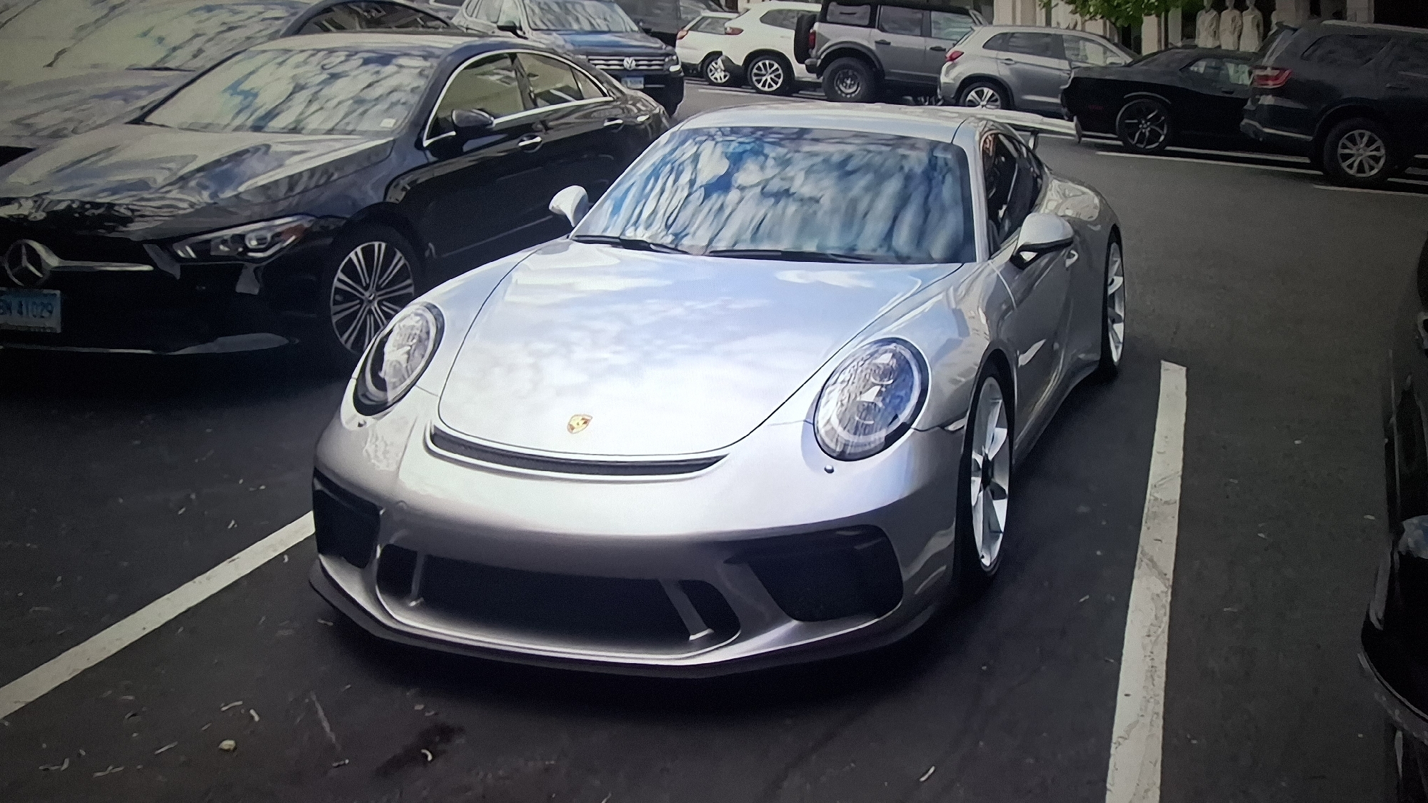 The image displays a daytime scene in an outdoor parking area, primarily featuring several parked automobiles. In the foreground, a silver Porsche 911 GT3 is prominently parked, viewed from a front-quarter angle. The car exhibits a sleek design with distinct front air intakes and the Porsche crest on its hood. Its highly reflective surface clearly mirrors the bright sky and cloud formations overhead. The car is fitted with silver multi-spoke wheels.
Immediately to the left of the Porsche, a black Mercedes-Benz sedan, identifiable by its front-end styling and badge, is parked. Part of its front bumper, headlight, and wheel are visible. A blue license plate with white characters "BN 41029" is attached to the Mercedes' front.
The background and surrounding areas show an assortment of other vehicles. These include a dark-colored SUV parked behind the Mercedes, and further back, a black muscle car resembling a Dodge Challenger, along with various other sedans and SUVs of different colors. Reflections on the windshields of these cars also suggest bright, possibly partly cloudy conditions.
The setting consists of an asphalt ground with white painted parking lines, indicating a parking lot or street parking. In the distant background, some light-colored architectural structures or building facades are faintly visible. No individuals are present in the frame, depicting a static scene of parked cars under clear or partly cloudy skies.