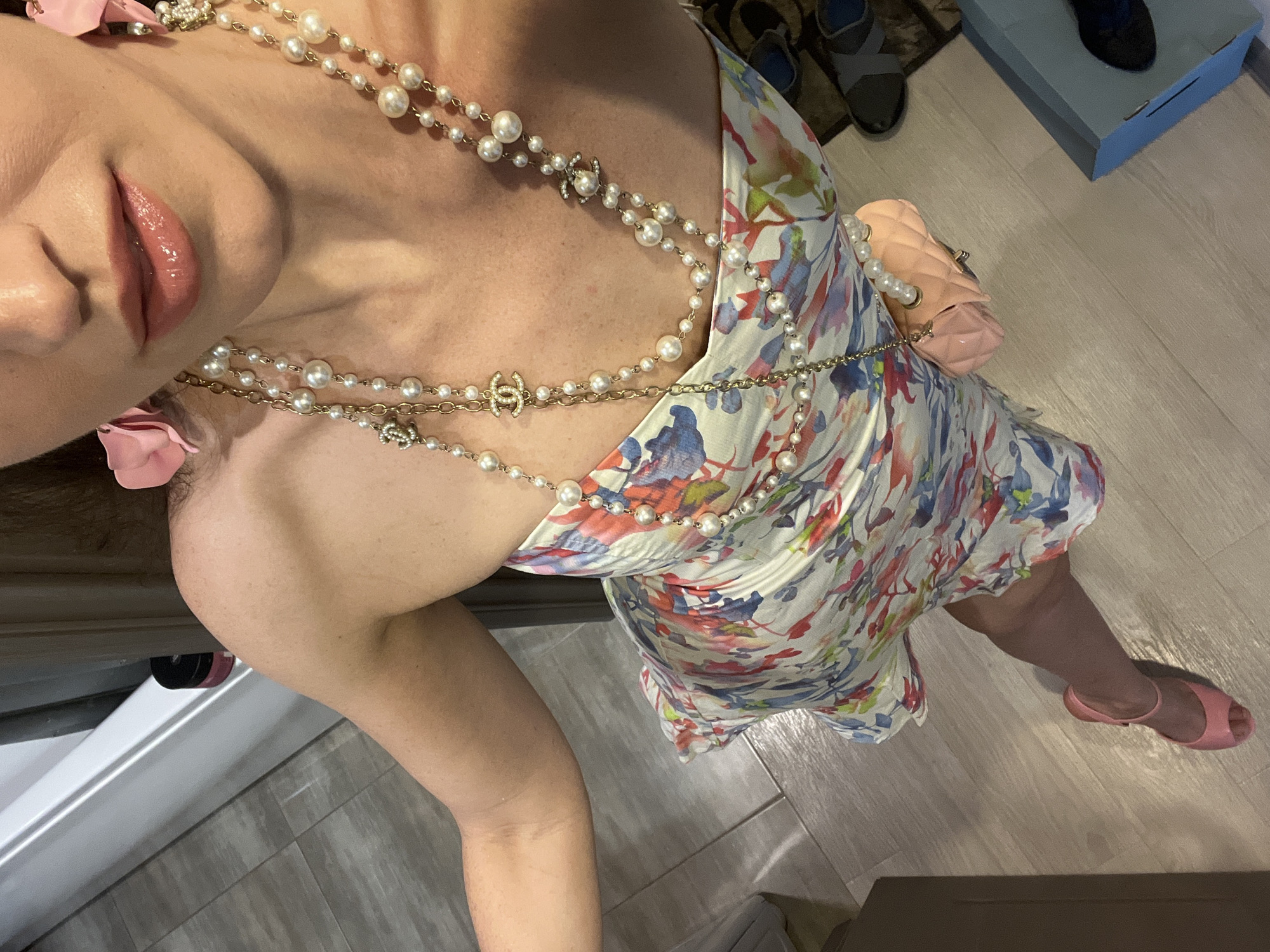 A woman is standing in a room with light-colored wood-look flooring. She is wearing a strapless dress with a floral print in shades of blue, pink, and red on a white background. She has a multi-strand pearl necklace with gold chain accents and Chanel logo charms. She is also wearing pink flower-shaped earrings and pink open-toed heels. She is holding a small, quilted pink handbag. The background shows a white baseboard and what appears to be a doorway or closet. There are some shoes visible on the floor to the right. The scene is indoors and does not provide any specific context or indicators related to the city of Kazan, Russia.