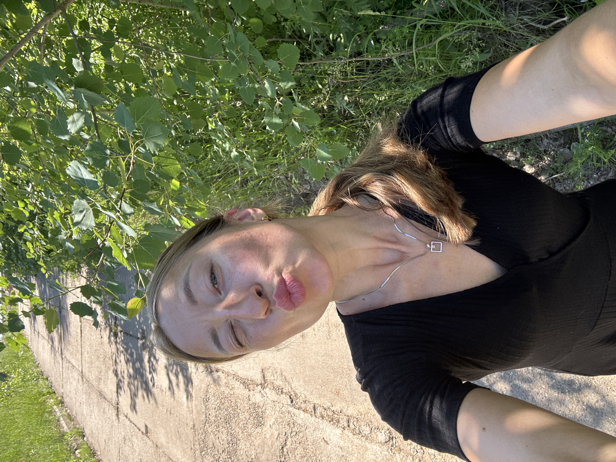 A woman with blonde hair in a ponytail, wearing a black dress and a silver necklace, poses for a selfie outdoors. She is winking and puckering her lips. The background consists of a paved path and lush green foliage, indicative of a park or natural setting. The image appears to have been taken in Chaikovsky, Russia.