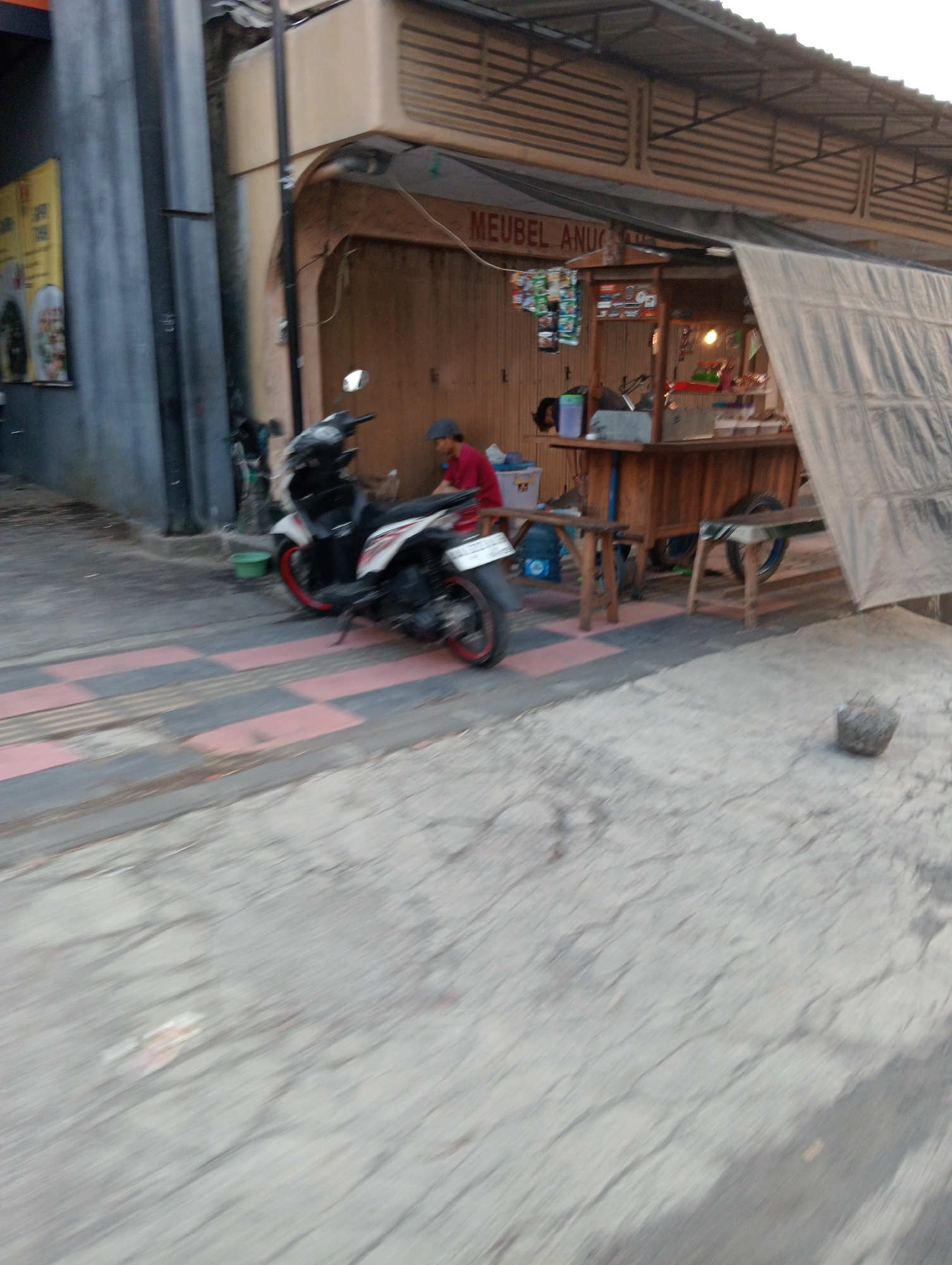 In Grabag, Indonesia, a street scene unfolds with a motorcycle parked on a checkered pavement in front of a small food stall. The stall, made of wood, has a tarp covering its roof, extending over the counter where food items and beverages are displayed. A person wearing a red shirt and a cap is seated at the stall. Behind the stall, a wooden building with a corrugated metal roof is visible, displaying a sign that reads "MEUBEL ANUGRAH." To the left of the stall, a wall with a yellow poster can be seen. The overall environment suggests an urban setting with a commercial activity taking place on the street.