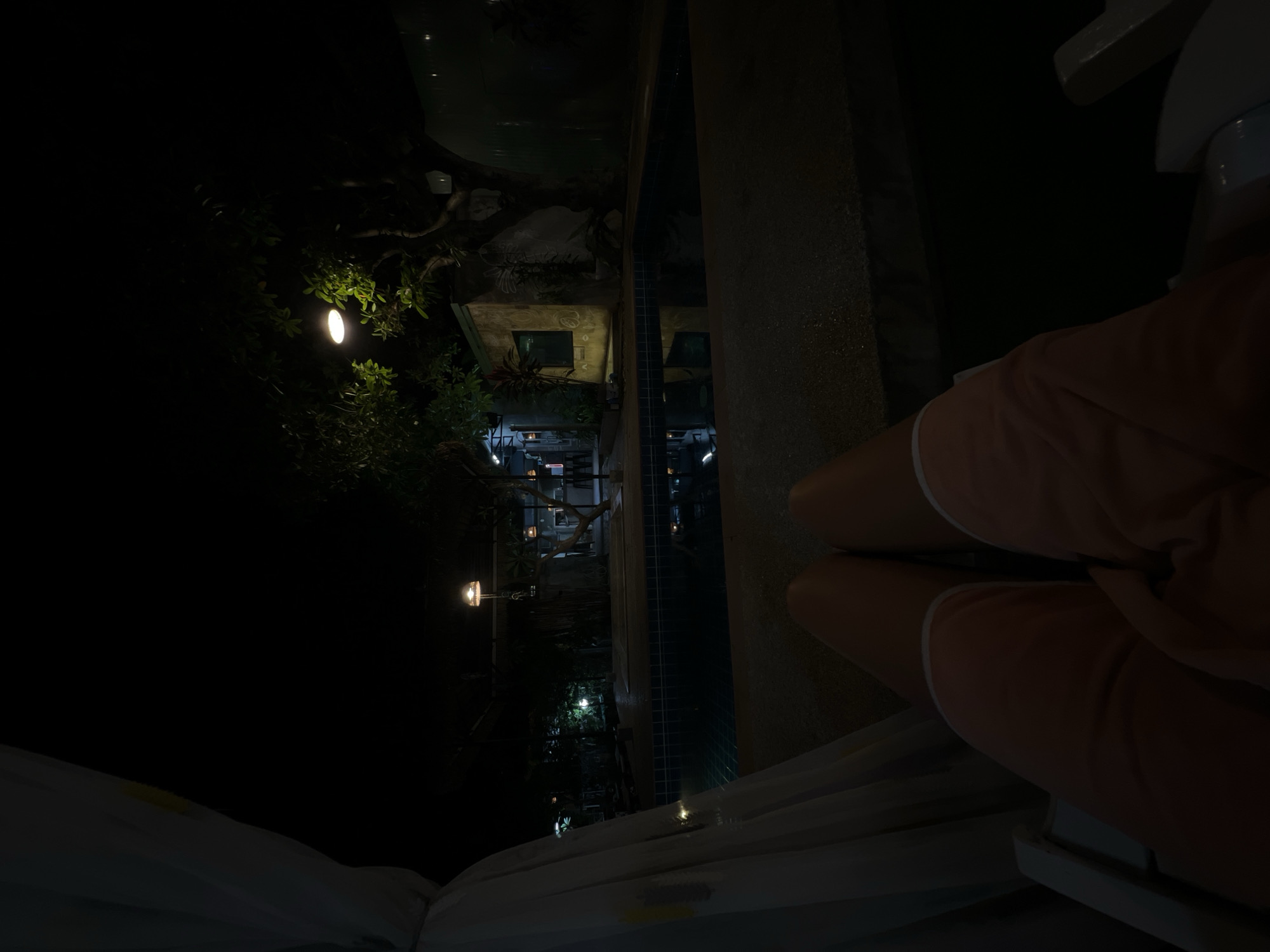 The image depicts a nighttime scene, possibly outdoors, with a person's legs visible in the foreground, wearing light-colored shorts with white trim. The person is sitting on what appears to be a white chair or lounger, with their knees bent. The setting seems to be near a swimming pool or body of water, with reflections visible on the surface. In the background, there are trees and foliage, illuminated by several small, warm-toned lights, creating an atmospheric and tranquil ambiance. A building with windows is partially visible, suggesting it might be a hotel, resort, or residential area. The lighting is dim, characteristic of nighttime, and the overall mood is serene and relaxed. There is no visible text in the image.