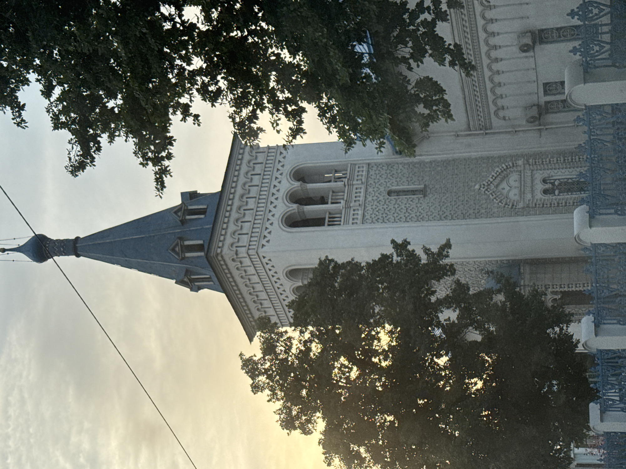 The image captures a distinct architectural structure, likely a church or cathedral, partially framed by lush green trees against a bright, cloudy sky. The building stands tall with a prominent blue, conical spire topped with an onion dome and a small finial or cross, dominating the left side of the frame. Its facade is a light, possibly white or cream, color, adorned with intricate decorative elements. These include repeated patterns of arches, geometric motifs, and detailed cornices along its upper sections and around windows. A section resembling a bell tower features multiple arched openings. Smaller, rectangular windows are also visible.
Dense foliage from trees, silhouetted against the bright sky, extends across the top and right side of the image, obscuring parts of the building and adding depth to the foreground. Sunlight filters softly through the clouds and tree branches, suggesting either early morning or late afternoon. A thin utility line or wire stretches diagonally across the upper left portion of the sky. On the far right, a segment of an ornate, blue metal fence or railing is visible, harmonizing with the spire's color. The scene is static, showcasing the building's grandeur and its interaction with the natural surroundings under a diffused light. Given the location context, the architecture is consistent with the Holy Resurrection Cathedral in Bishkek City, Kyrgyzstan.