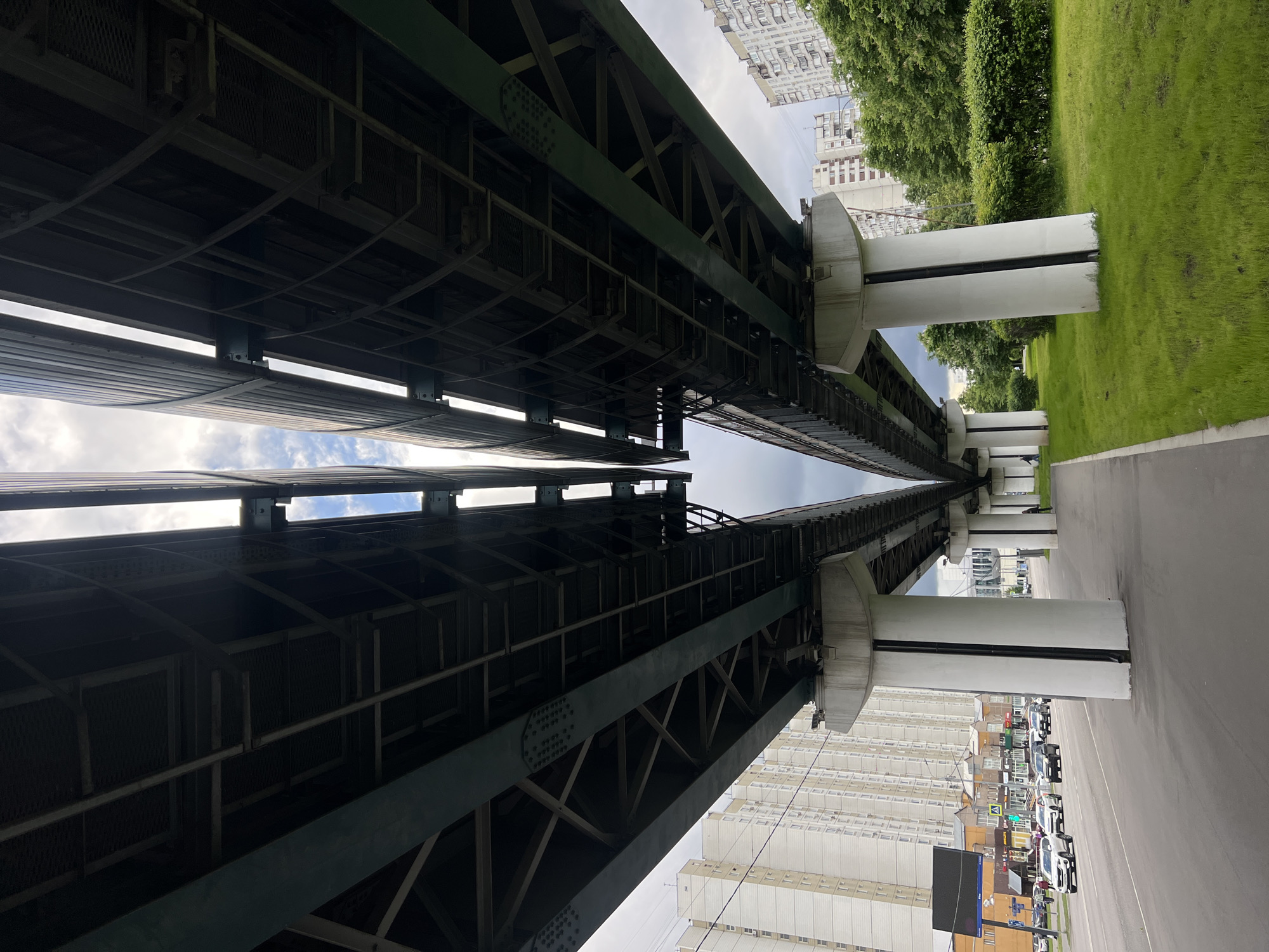 The image depicts an elevated railway structure in Moscow, Russia. The view is from below, looking up at the tracks and the supporting pillars. The structural steel is painted green and is intricate, with many crossbeams and lattice sections. The pillars supporting the structure are white and rectangular. Adjacent to the railway are buildings, a grassy area with trees, and a roadway with parked cars. The sky is visible between the tracks, with a cloudy atmosphere.