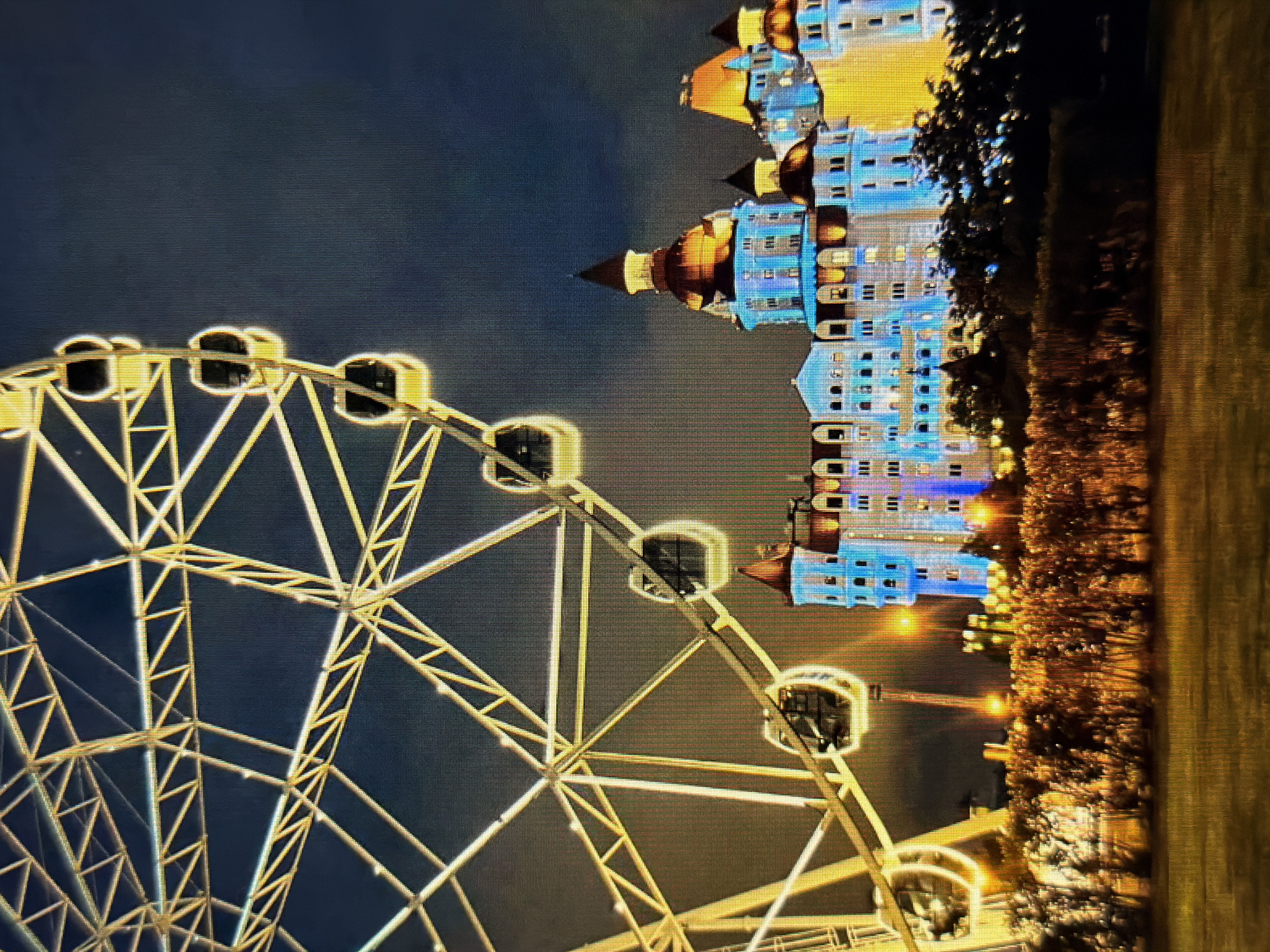 The image shows a nighttime scene in the city of Sirius, Russia. A large, brightly lit Ferris wheel dominates the left side of the frame. The Ferris wheel is yellow and white, with several gondolas visible, each illuminated with a soft glow. To the right of the Ferris wheel, a multi-story building is illuminated with blue and yellow lights, featuring a distinctive domed roof. The building's architecture appears ornate, with many windows. In the foreground, there is a row of bushes or trees, also illuminated by artificial lights, creating a warm, orange glow. The ground in front of the bushes appears to be a grassy area. The sky is dark, indicating nighttime, with some cloud cover. The overall impression is of a vibrant and festive amusement park or entertainment complex.