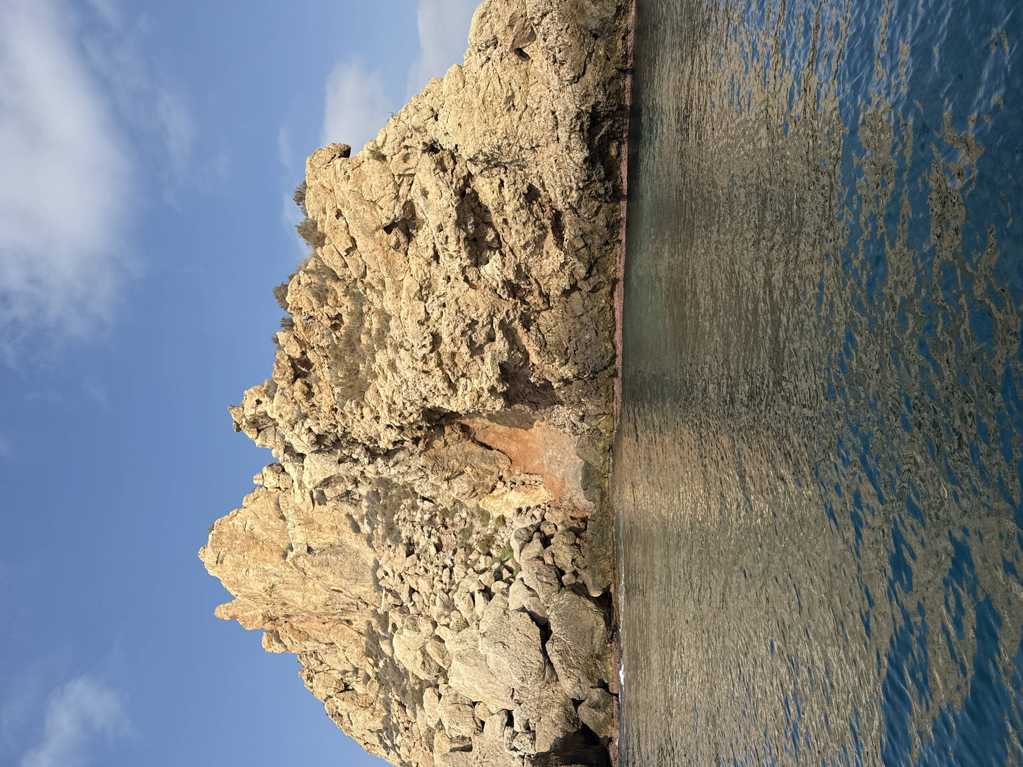 A large, weathered rock formation dominates the center of the frame, rising from the calm, dark blue water of the Mediterranean Sea. The rock is a light tan color, with many sharp edges and crevices, indicating significant erosion. There are sparse patches of dry, brown vegetation clinging to the upper slopes. The lower portion of the rock is dark and wet, suggesting it is submerged at high tide. The water in the foreground is clear, with a slight ripple, and reflects the sunlight and the colors of the rock. The sky above is a clear, bright blue with a few wispy white clouds. This rock formation is located in the vicinity of Cabo Cope, a protected natural area near the city of Águilas, Spain.