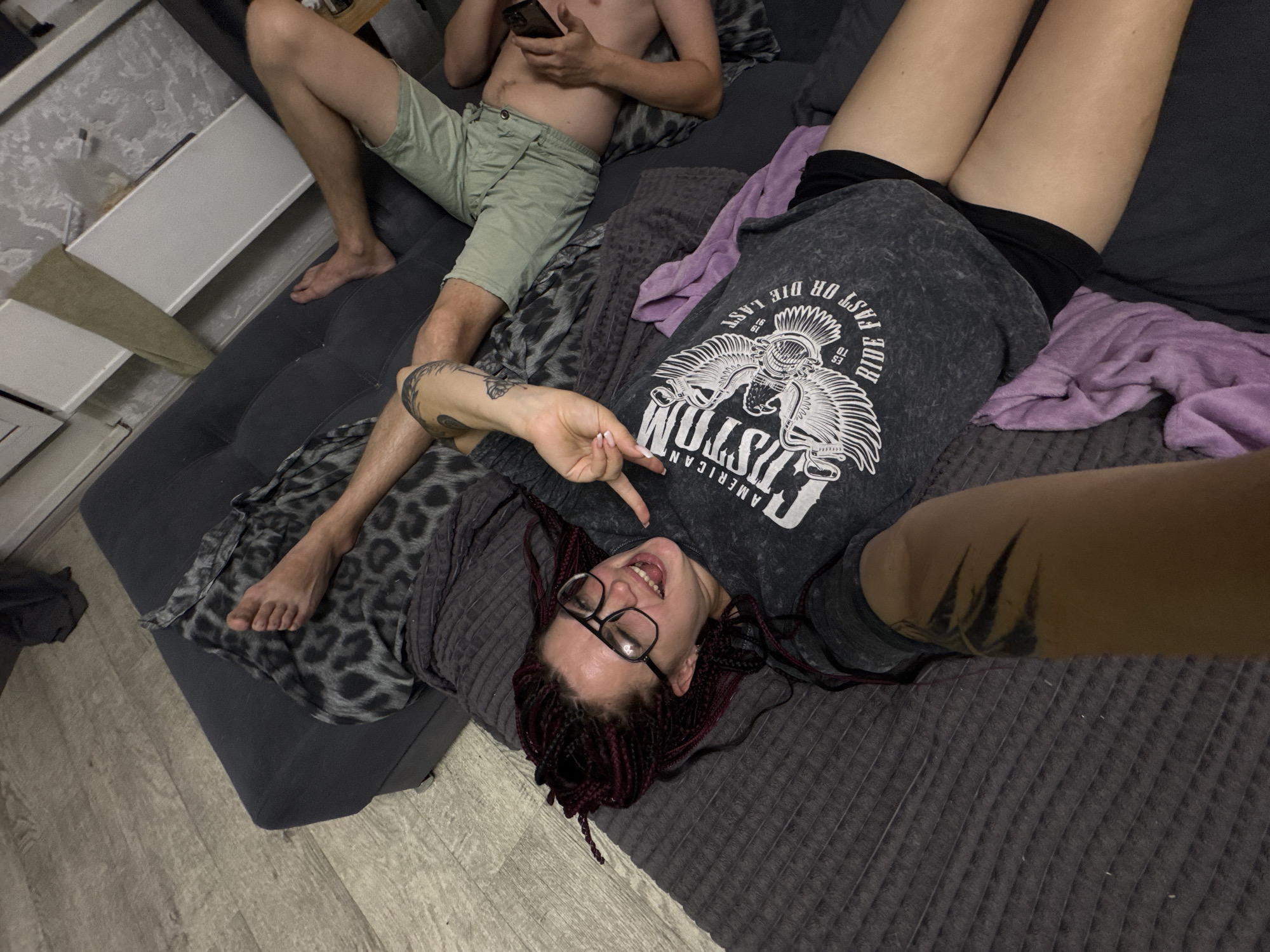 The image shows two people on a couch in a dimly lit room. One person, with light skin and light brown hair, is lying on their back, looking up at the camera. They are wearing glasses, a gray t-shirt with a graphic print, and black shorts. Their hair is styled in dark red braids. They are smiling and making a peace sign with their right hand. Tattoos are visible on their left arm.
The other person is lying on their side, partially visible in the upper left of the frame. They have light skin and are wearing light green shorts and no shirt. They are holding a phone in their right hand.
The couch appears to be a dark gray sectional, covered with a dark gray patterned blanket. A purple blanket is draped over the couch. The floor is made of light-colored wood-look laminate. In the background, on the left, there is a white cabinet or shelf unit.
The scene appears to be indoors, likely a living room, at night or in a room with minimal natural light. The mood is casual and relaxed. The t-shirt the person in the foreground is wearing has text that includes "AMERICAN CUSTOM" and "RIDE FAST OR DIE LAST". The overall impression is one of a relaxed, informal moment between two individuals.