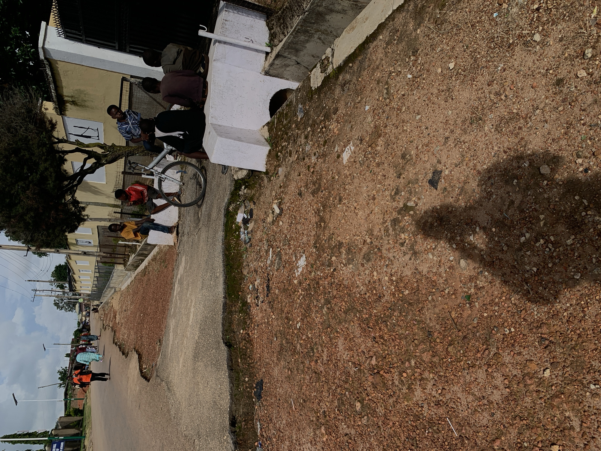 A group of young Black males are gathered near a concrete structure and a bicycle on a dirt road. Some are sitting on the concrete, while one is seated on the bicycle. Another individual, wearing a blue and white checkered shirt, is standing and looking at the seated group. In the background, several people walk along a paved road that runs alongside the dirt area. The scene appears to be a street or pathway in a community, with buildings and trees visible. The weather is overcast.