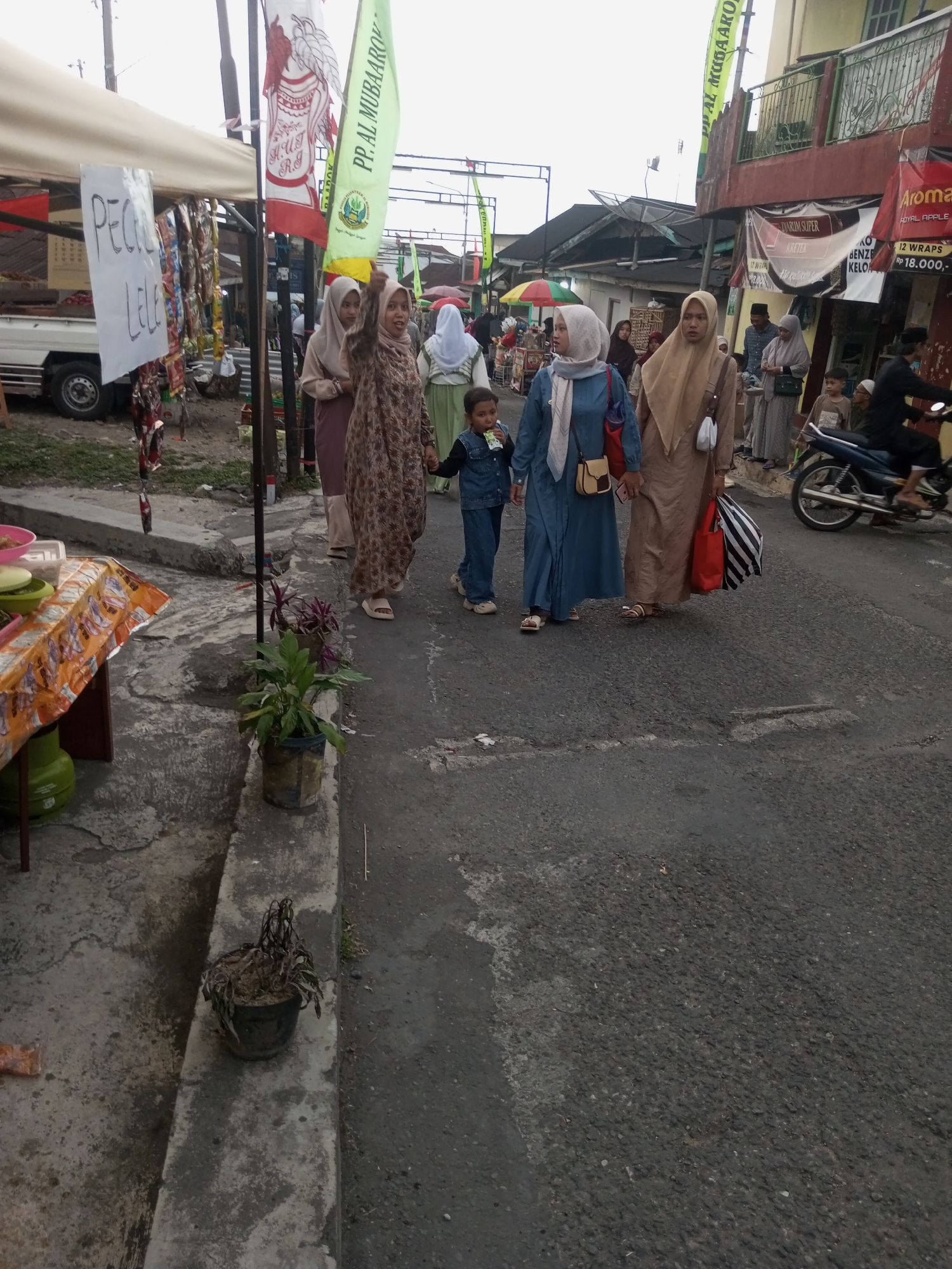 In Pandansari, Indonesia, four individuals are walking on a paved road lined with market stalls. Three women are dressed in hijabs and long dresses. The woman on the far left wears a patterned brown dress, the woman next to her wears a blue dress, and the woman on the far right wears a tan dress. A young child in a denim outfit is walking between the woman in the blue dress and the woman in the tan dress, holding their hands. In the background, several market stalls are visible, some with awnings and displays of goods. There are also buildings with balconies and a motorcycle being ridden by a person in dark clothing. The environment appears to be a bustling market scene.