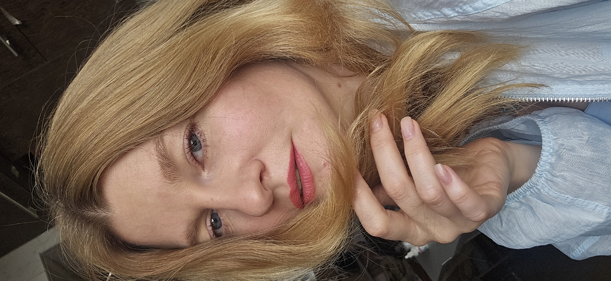 A woman with blonde hair and blue eyes is shown from the chest up, holding a strand of her hair with her right hand. She is wearing a light blue blouse. Her lips are painted with a coral lipstick. The background is dark and out of focus. The image was taken indoors.