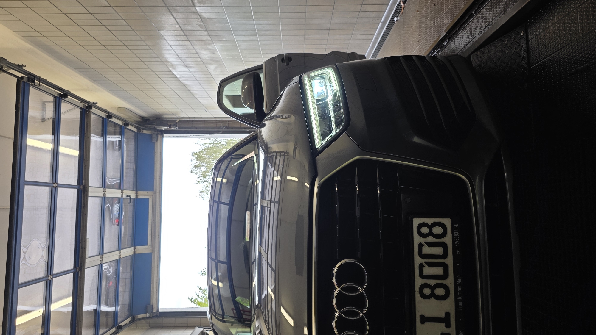A dark gray Audi Q3 SUV is parked inside a car wash bay with the driver's side door open. The car faces forward, with its headlights illuminated. The bay has a tiled wall on the right and a large, segmented garage door with clear panels at the top. Through the open garage door, trees and a cloudy sky are visible. The car has a license plate with "I 8008" and "Frankfurt am Main" printed on it. The floor of the bay is a dark, patterned metal surface, and the car is elevated slightly on a raised platform.