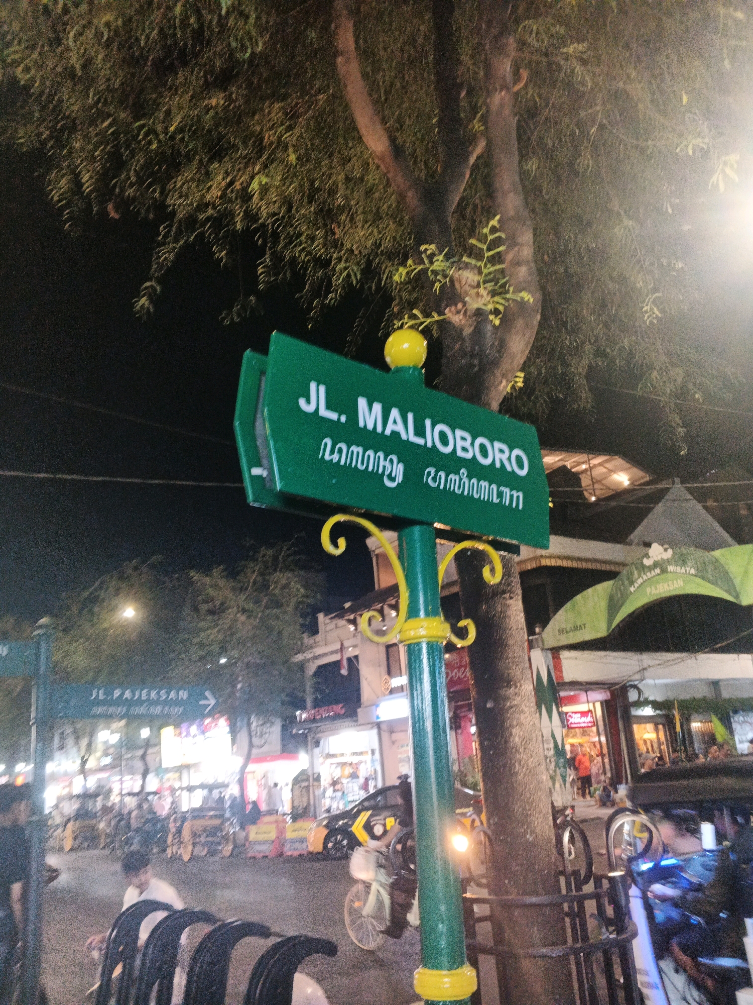 The image depicts a street scene at night in Yogyakarta, Indonesia, specifically focusing on a street sign that reads "JL. MALIOBORO." The sign is green with white lettering and features an inscription in Javanese script below the main text. A yellow spherical finial sits atop the signpost. The signpost itself is green with yellow decorative elements.
Behind the sign, a large tree trunk is visible, with dense foliage overhead partially obscuring the night sky. Streetlights and the lights from surrounding buildings cast a warm glow on the scene.
In the background, the street is bustling with activity. Several horse-drawn carriages, known as "andong," are parked or moving, indicating a tourist area. There are also various shops and stalls lining the street, some with illuminated signs. People are visible walking and interacting, suggesting a lively night market or pedestrian zone. Another street sign is visible in the background, pointing to "JL. PAJEKSAN." The overall atmosphere is that of a vibrant urban environment at night, characteristic of a popular tourist destination.