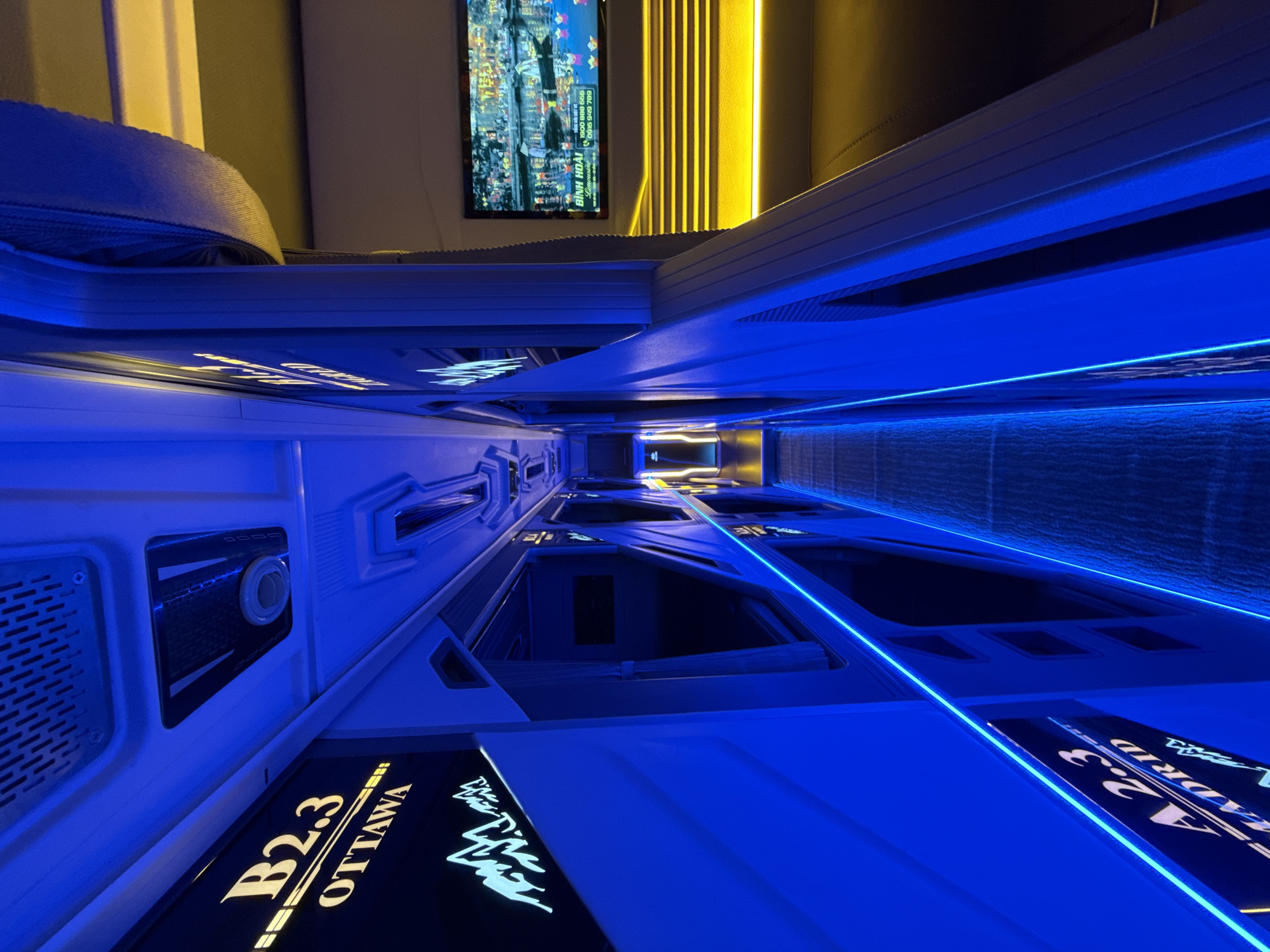 The image displays the interior of a modern, futuristic capsule sleeping facility, characteristic of a capsule hotel or a premium sleeping bus/train, set in Đà Nẵng, Vietnam. The scene is illuminated by striking electric blue LED strip lighting that lines the edges of multiple individual sleeping pods and horizontal surfaces, creating a high-tech atmosphere.
The perspective looks down a narrow corridor or aisle, flanked by rows of identical, sleek capsule compartments on both sides. Each capsule features a dark, recessed opening. On the side panels of the pods, illuminated text provides specific identifiers and thematic names, such as "B2.3 OTTAWA" and a partially visible "A 2.3 MADRID."
At the far end of the corridor, bright yellow-orange vertical strip lights offer a contrasting glow. Above these lights, a vertical screen or monitor displays a vibrant image of a city skyline at night. Text visible on this screen includes Vietnamese words "BÌNH HOÀI," phone numbers "1900 888 666" and "0936 949 789," and the word "Limousine."
No people are present, and no specific activity is taking place; the scene is static, showcasing the accommodation. A rolled-up grey fabric, likely bedding, is visible inside one of the capsules in the foreground. One capsule interior features a small circular vent or control panel. The artificial lighting suggests an interior setting, contributing to a modern, almost sci-fi ambiance.
