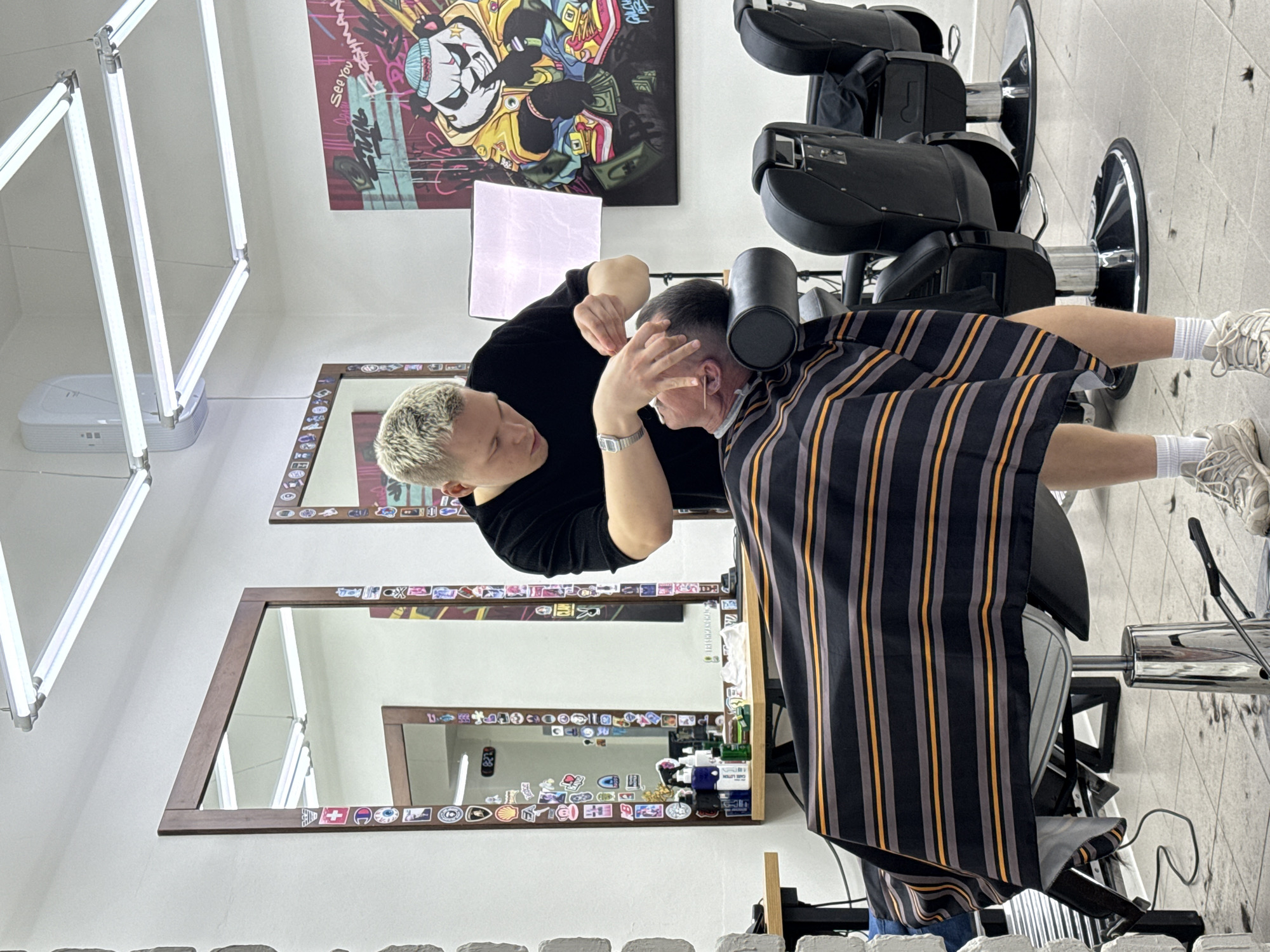 The image depicts a barber giving a haircut to a customer in a barbershop. The barber has short, bleached blonde hair and wears a black shirt. The customer is sitting in a barber's chair with a black and gold striped cape draped over him. The barber is using his hands to style the customer's hair.
The environment appears to be a modern barbershop with two mirrors framed with stickers. There is a bright lighting fixture on the ceiling and artwork on the wall.
Based on the prompt, the location is a city of Chaikovsky, Russia.