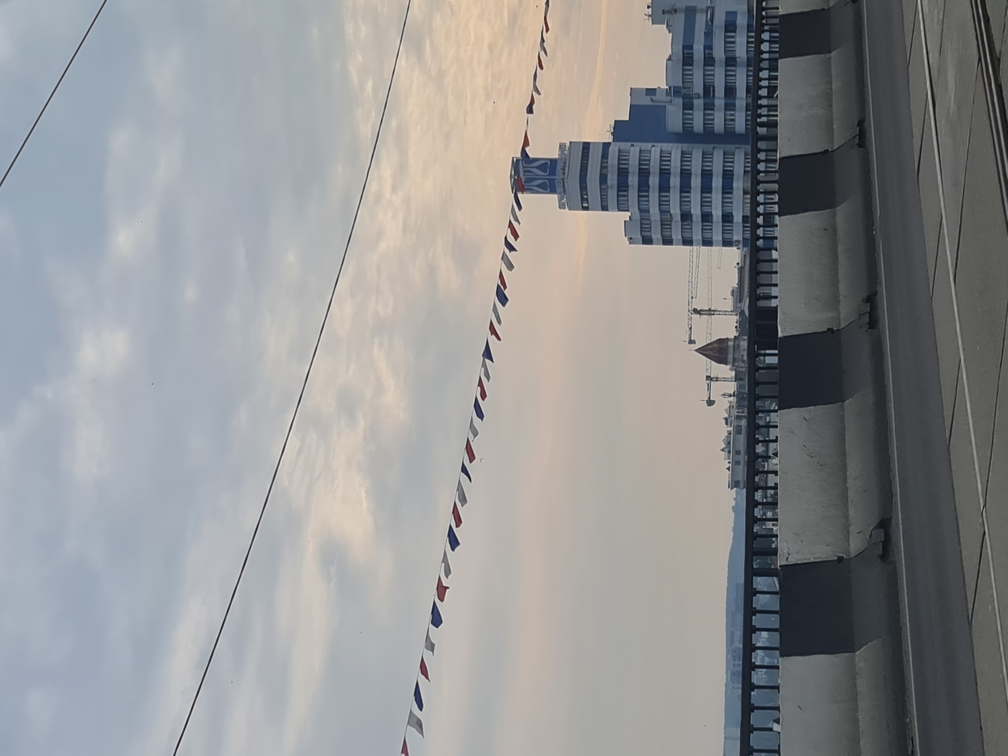 This image depicts a daytime view from a moving vehicle on a bridge in Irkutsk, Russia. The foreground shows a section of the bridge's roadway and a concrete barrier with a black and white pattern. In the mid-ground, a railing runs along the edge of the bridge, behind which a city skyline is visible. Dominating the skyline is a tall, multi-story building with blue and white accents, topped with a distinctive spire-like structure. In the background, clouds fill the sky, with power lines stretching across the frame. A string of small, triangular flags in red, white, and blue is draped across the sky, suggesting a festive occasion or decoration. Construction cranes are also visible in the distance, indicating ongoing development.