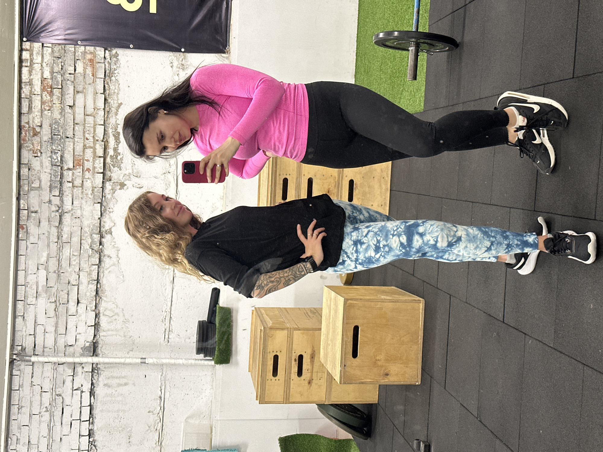This image captures two women posing in an indoor gym setting in Chaikovsky, Russia.
On the left, a woman with dark hair wears a bright pink long-sleeved top, black leggings, and black and white athletic shoes. She holds a red smartphone at chest height, seemingly taking a selfie or recording a video. Her expression is neutral, looking towards the phone.
On the right, a woman with wavy blonde hair wears a black long-sleeved shirt, blue and white tie-dye patterned leggings, and black and white athletic shoes. She has visible tattoos on her left forearm. She also looks towards the phone with a relaxed, slight smile.
The setting is a fitness studio with a dark grey interlocking mat floor. The background includes a white brick wall on the left and a plain white wall. Several wooden plyo boxes are stacked behind the women, and a barbell weight plate on a rack is partially visible in the upper right. Small patches of green artificial turf are seen near the equipment. A black banner hangs at the top, displaying a partial yellow character that resembles the number "8." The lighting suggests it is daytime.