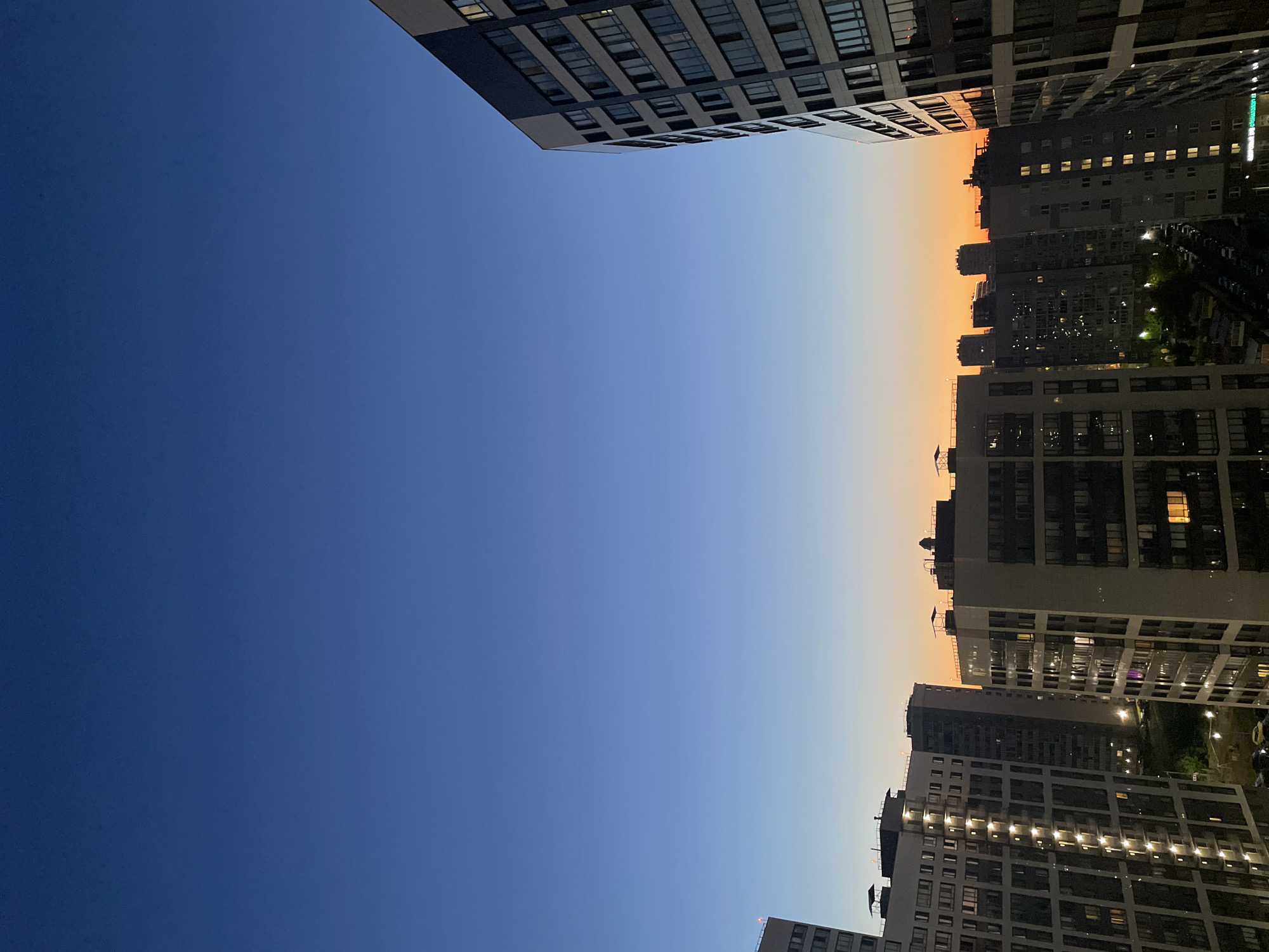 The image depicts a twilight view of a city skyline, possibly Sinyavino, Russia, characterized by multiple tall apartment buildings. The sky transitions from a deep blue at the top to a warm orange hue near the horizon, indicating either sunrise or sunset. The buildings are lit with numerous windows, suggesting residential occupancy. Some structures are modern, with sleek facades and repetitive window patterns, while others appear older and more utilitarian. The foreground features a prominent, modern apartment building with a dark, grey facade and numerous windows. In the mid-ground and background, other apartment complexes are visible, some with balconies. The lower parts of the buildings show street-level details like parked cars and streetlights. The overall scene is an urban landscape captured during the transitional light of dawn or dusk. There are no people or animals visible in the image.