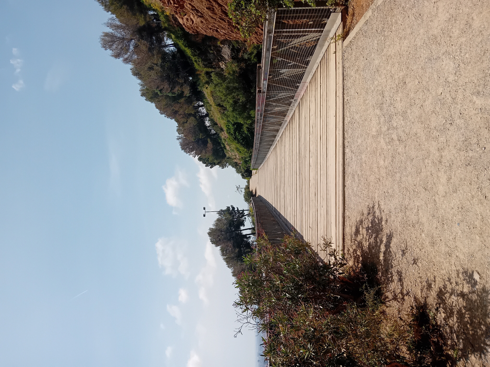 A scenic outdoor pathway, likely a promenade or trail, is depicted under a bright, clear blue sky. The image features a wide, light-colored wooden boardwalk running alongside a parallel dirt or gravel path. A metal mesh fence with a top railing separates the boardwalk from a steep, green hillside on the left, which exhibits reddish-brown rock formations near its crest. The hillside is densely covered with trees and shrubs, typical of Mediterranean vegetation.
In the foreground, particularly on the bottom right, lush green bushes with some reddish foliage cast long shadows across the gravel path, indicating a sunny day with the sun at a moderate angle. A few streetlights are visible further down the pathway in the mid-distance. The sky, occupying the upper portion of the image, is mostly clear with a few scattered white clouds, and a faint airplane contrail can be seen in the upper left.
No people or specific activities are captured, but the path appears suitable for walking or leisure. There are faint, unidentifiable markings resembling graffiti on the metal fence. The overall atmosphere is peaceful and serene, suggesting a pleasant outdoor environment. The setting, with its specific vegetation and rocky hillside bordering a constructed walkway, is consistent with a coastal or hillside trail found in Barcelona, Spain.