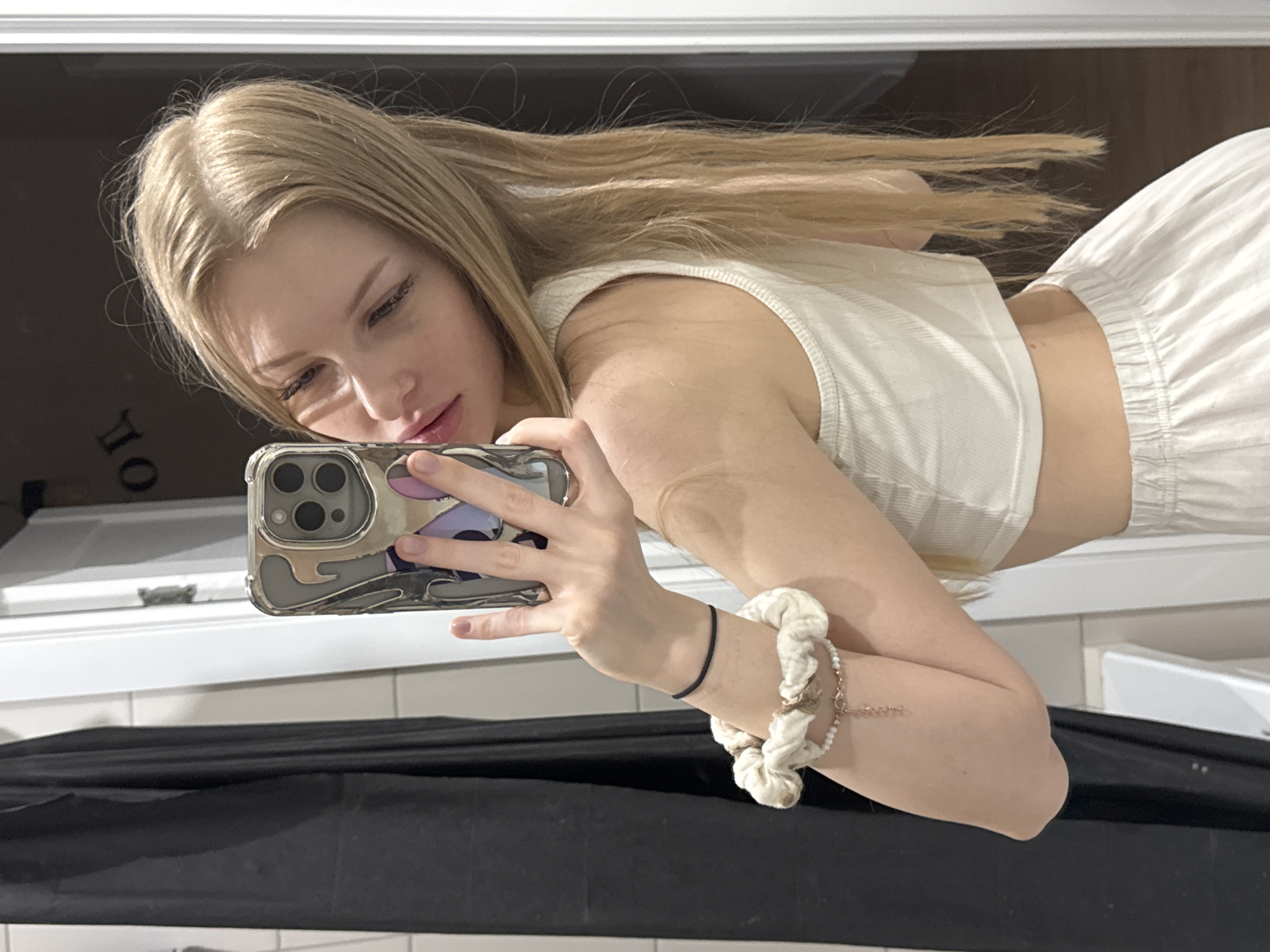A young, light-skinned female with blonde hair is taking a selfie in what appears to be a bathroom. She is wearing a white crop top and white sweatpants. She is holding a phone with a grey case. The phone is positioned to capture her face and torso. On her left wrist, she wears a black elastic hair tie and a white scrunchie with a delicate rose gold beaded bracelet. The background shows a white countertop, a black material draped in front of it, and a blurred reflection in a mirror. The location is not discernible as being in Perm, Russia from the available visual information.