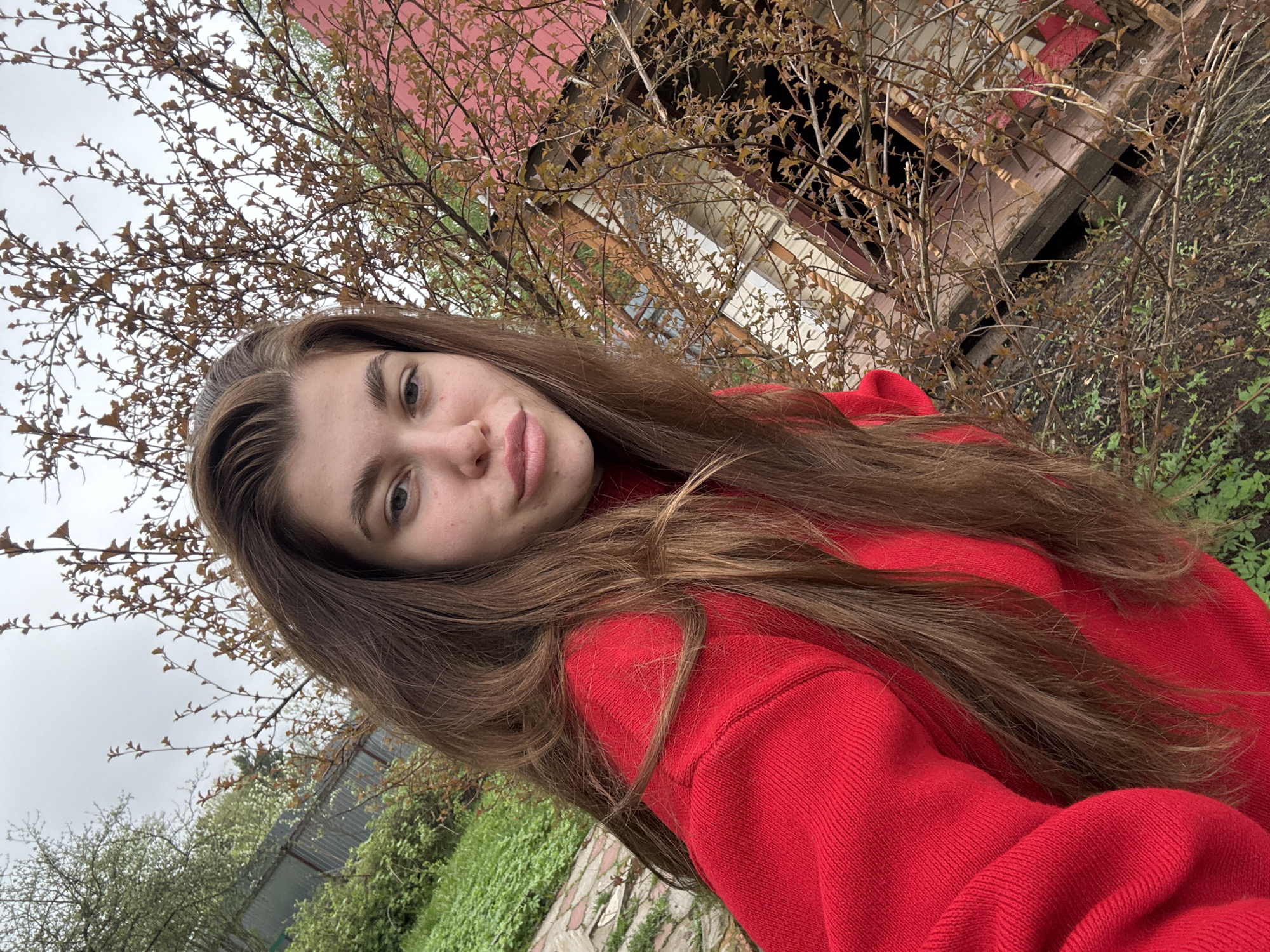 A young woman with long brown hair stands outdoors, wearing a red sweater. She is facing the camera with a slight smile. Behind her is a weathered, possibly abandoned, wooden structure with a red roof, partially obscured by branches. Green foliage and a cloudy sky are also visible in the background. The location could be in a rural area.