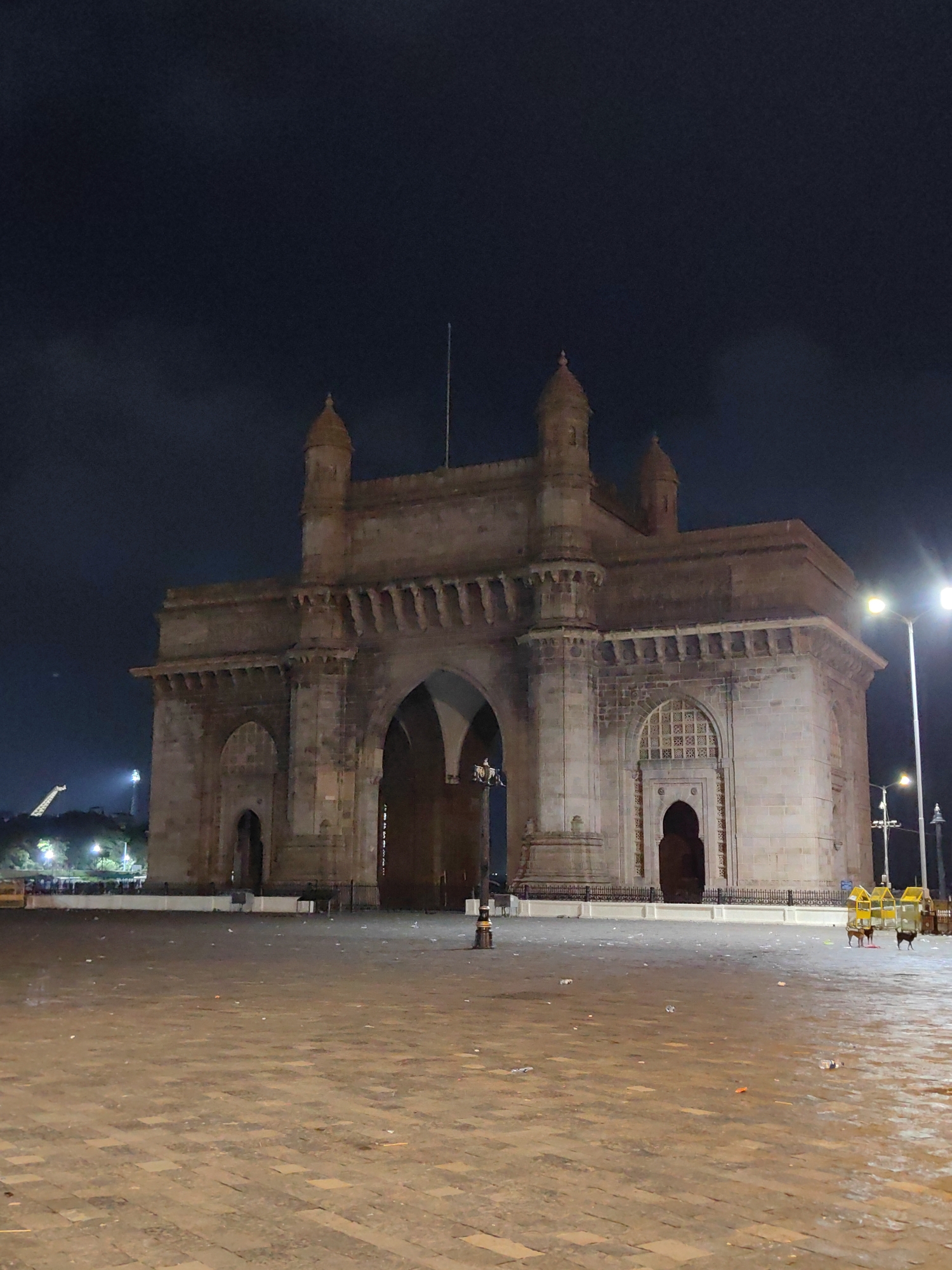 The image shows the Gateway of India monument in Mumbai, India, at night. The monument, constructed of red basalt, is illuminated by streetlights and appears to be made of stone blocks. It features a large central arch and two smaller arches on either side. The area in front of the monument is a paved plaza, littered with scattered debris, including plastic bottles and wrappers. Two yellow trash bins are visible on the right side of the plaza. In the background, on the left, a tall structure resembling a giraffe is visible, along with trees and more distant lights. The sky is dark and cloudy. No people are clearly visible in the immediate vicinity of the monument.