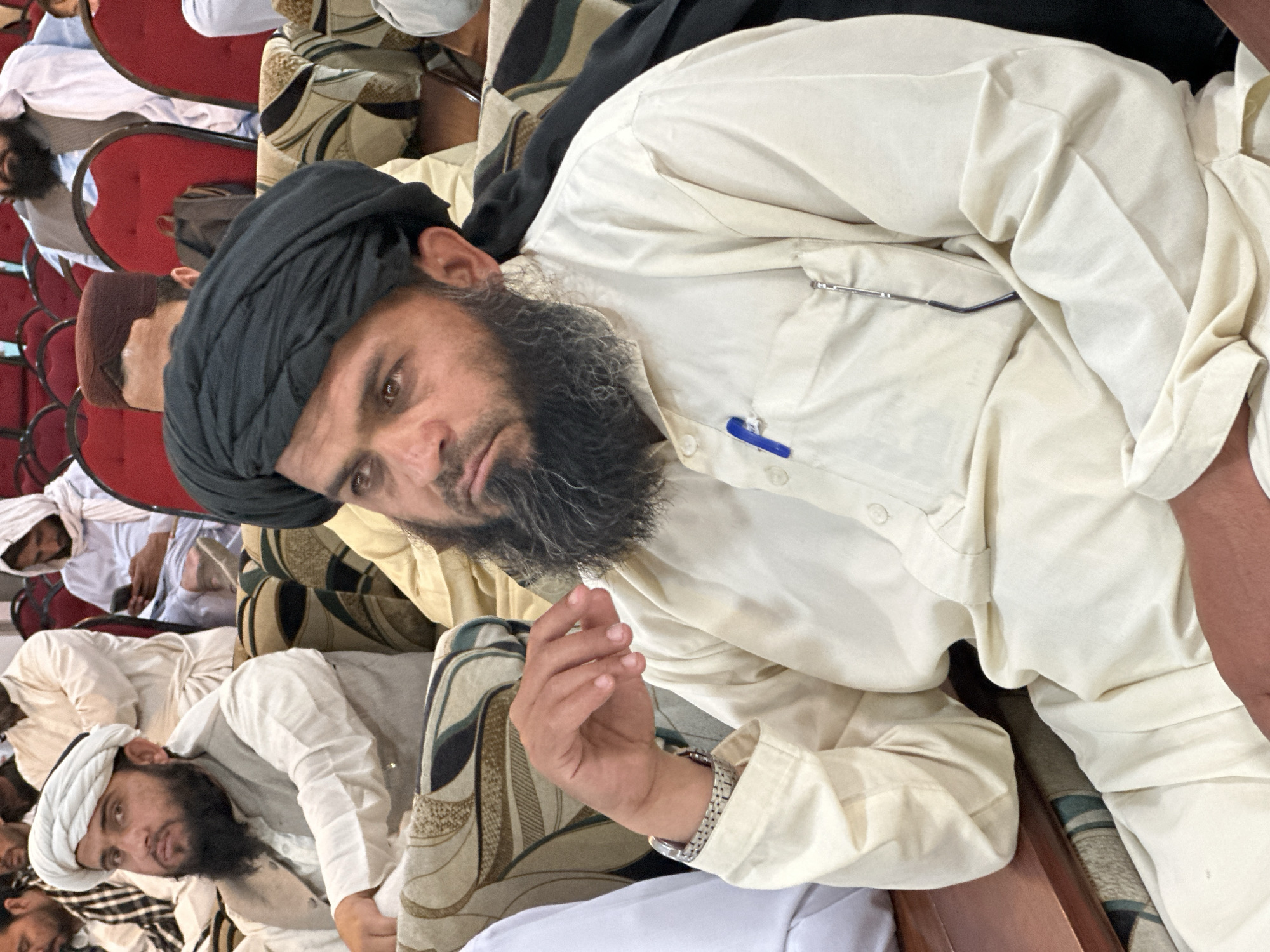 The image shows a man with a dark beard and a black turban, wearing a light-colored shirt with a pen clipped to the pocket. He is seated among other men, some of whom are wearing similar attire. The environment appears to be indoors, possibly a meeting or gathering. Based on the prompt specifying the city of Quetta City Tehsil, Pakistan, it's inferred the image was taken in that location.
