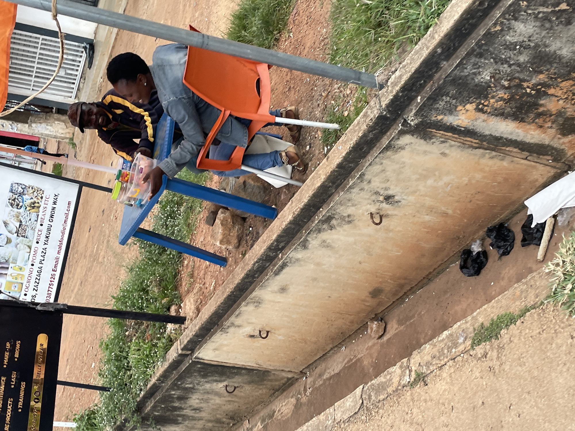 The image depicts an outdoor scene, likely in Jos, Nigeria, with a focus on a street-level environment featuring a concrete drainage channel and two individuals seated at a table.
In the foreground, a substantial concrete drainage channel runs along the left side of the frame. Its interior appears damp and somewhat dirty, with visible debris including several black plastic bags, some of which contain what looks like trash. A few rocks and patches of grass are scattered within and around the channel.
In the mid-ground, two people are seated at a small, bright blue table. A man wearing a dark jacket with yellow stripes and a brown baseball cap is seated on the left, facing the woman. He appears to be smiling. The woman on the right is wearing a denim jacket and blue jeans. She is holding a clear plastic container filled with food items, seemingly engaging in a conversation with the man. An orange plastic chair is positioned to her right, supporting her. The table itself has a few items on it, including the food container and what appear to be jars or bottles.
Behind the table, a dark sign with text and logos is partially visible, along with metal poles supporting an orange awning that extends across the top right portion of the image. The background shows a reddish-brown dirt road or ground, with some vegetation and scattered rocks. A person in a blue shirt is walking in the distance. The overall impression is a casual, everyday scene at a roadside vendor or gathering spot in Jos.