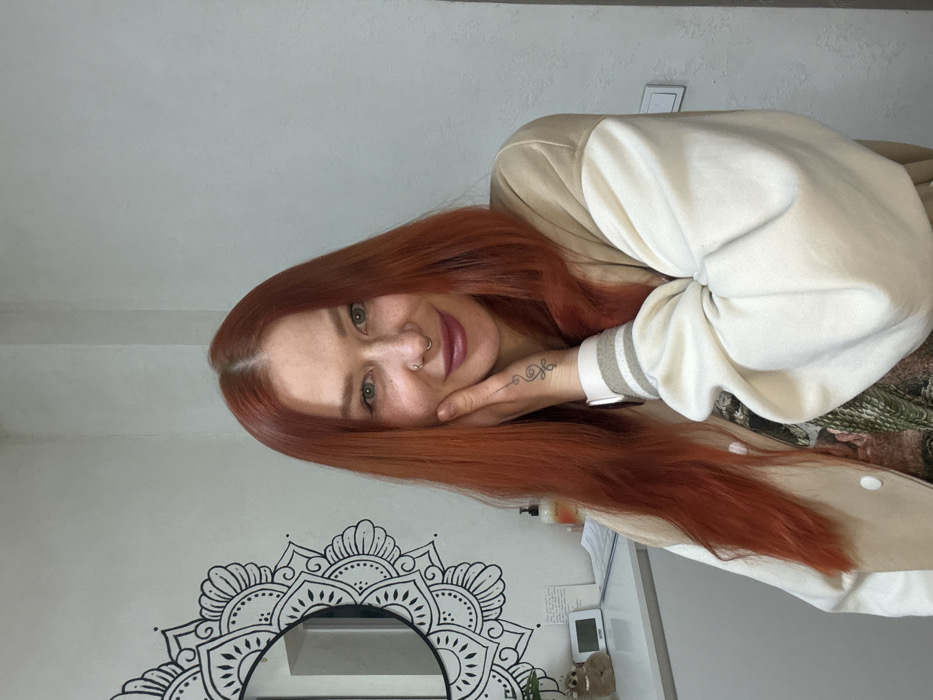 A woman with red hair and green eyes is smiling with her face resting on her hand. She's wearing a light-colored varsity jacket. A black and white mandala design decorates the wall behind her. Also visible are a mirror, a small figurine, and what appears to be a thermostat or timer. The photo is taken from a low angle, making the woman appear to look down at the viewer.