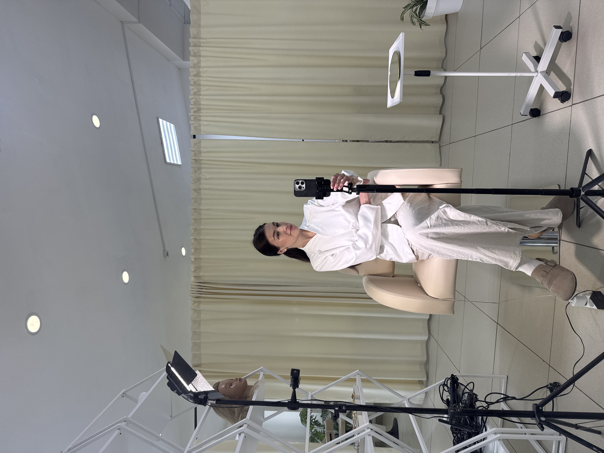 An indoor scene shows a woman with dark hair, wearing a white shirt or robe and light-colored pants, seated in a beige armchair. She is holding a black smartphone, mounted on a small tripod, in front of her, suggesting she is recording or participating in a video call. Her gaze is directed towards the camera of the phone.
The setting appears to be a well-lit studio or professional room, possibly in Chaikovsky, Russia, with white walls, a light-tiled floor, and light beige curtains in the background. Several pieces of professional equipment are visible: a large white ring light or panel on a stand is positioned behind the woman to her right. In the foreground, another professional lighting setup is present, featuring a blonde mannequin head and a laptop with an illuminated screen, both positioned on a white, lattice-like structure that resembles a stand or shelf. Cables and power adapters are visible on the floor around this equipment.
The room is illuminated by multiple sources, including recessed circular lights in the ceiling and a rectangular overhead fluorescent light. A potted plant can be seen in the upper right background. The woman is wearing light-colored, possibly suede, slip-on shoes. The overall atmosphere suggests a planned recording session or online event, possibly a tutorial or product demonstration, within a clean and modern environment. No discernible text is visible in the image.