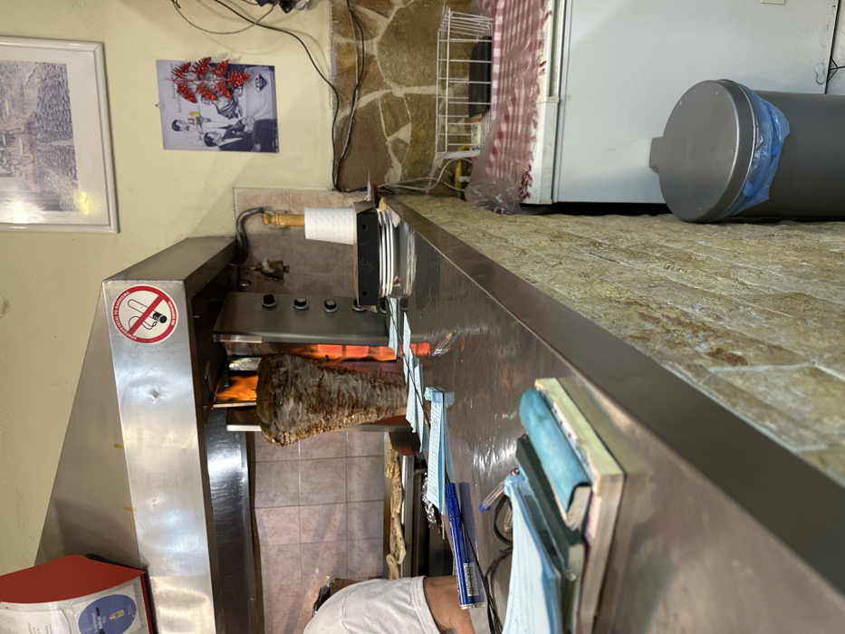 This image captures the interior of a bustling food preparation area, likely a Greek gyros or doner kebab shop in Dafni, Greece. The central focus is a large, stainless steel vertical rotisserie, actively cooking a sizable cone of meat over visible flames.
A red and white circular no-smoking sign is prominently displayed on the side of the rotisserie, featuring a crossed-out cigarette and the Greek text "ΑΠΑΓΟΡΕΥΕΤΑΙ ΤΟ ΚΑΠΝΙΖΕΙΝ," meaning "SMOKING IS FORBIDDEN." Above the cooking apparatus, a paper towel dispenser holds a fresh roll.
The background features walls partially painted a light beige and partially adorned with rustic stone-like tiles. A framed black and white picture hangs on the wall, depicting people, possibly enjoying drinks, embellished with red berry decorations. Another framed image with an unclear subject is seen further left. Cluttered wiring runs along the wall near a small white wire rack.
To the right, a higher counter or shelf is visible, surfaced with light-colored, textured tiles. On this surface, a white refrigerator and a grey cylindrical object, possibly a water heater or storage container with a blue bag on its lid, are present. In the lower right foreground, a person wearing a white t-shirt is partially visible, suggesting an employee is at work. The stainless steel counter in the immediate foreground holds various work-related items such as pens, notebooks, and papers. The scene conveys an active cooking environment.