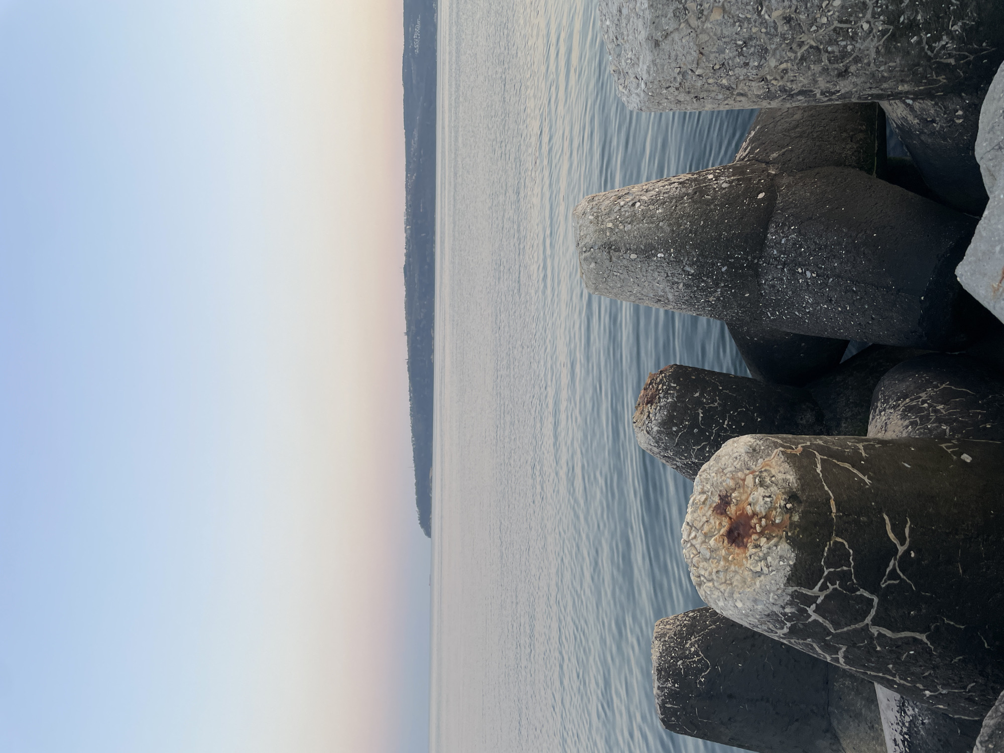 The image depicts a coastal scene, specifically a breakwater made of tetrapod concrete structures along the Black Sea coast in Varna, Bulgaria. The concrete tetrapods are stacked together, creating a barrier against the sea. Their surfaces are weathered, with visible cracks and discoloration, indicating exposure to the elements. In the background is the sea and a blurred line of land, which is likely the coast of Bulgaria. The sky transitions from a pale blue to a light orange, suggesting either sunrise or sunset. The water surface shows slight ripples, implying a calm sea state. No people are visible in the image.