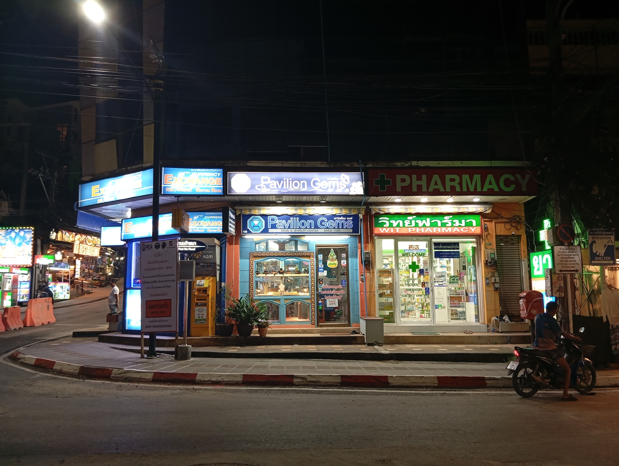 At night in Karon, Thailand, a street scene unfolds with a row of illuminated storefronts lining the curved roadway. To the left, a currency exchange booth displays a bright blue sign with "EXCHANGE" prominently featured. Next to it, a jewelry store named "Pavilion Gems" has a blue facade with a display window showcasing various items, and a sign indicating "GEMS & JEWELRY CENTER SINCE 1995". Further to the right, a pharmacy with a green awning and a red "PHARMACY" sign is visible, with its interior illuminated and stocked with products. Several people are present, including a man standing near a construction zone sign, and another man riding a motorcycle. The road is paved, with a red and white striped curb marking the edge of the sidewalk. The overall atmosphere is one of a commercial street at night, with buildings and signs lit up against the dark sky.