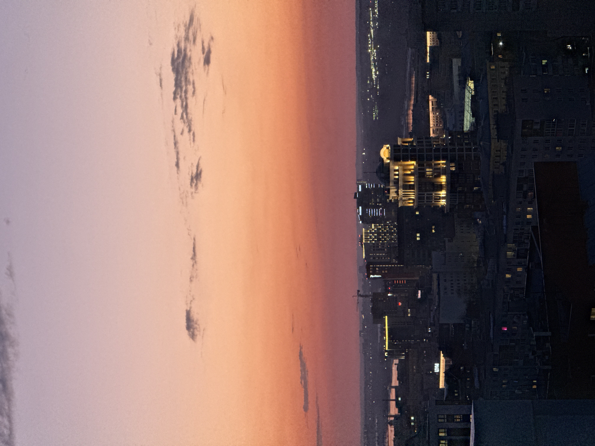 The image depicts a cityscape at dusk. The sky transitions from a light purple-gray on the left to a peach-orange hue towards the center. Scattered clouds are visible in the sky. The city of Perm, Russia, is on the right side of the image and illuminated with artificial lights. A mix of tall buildings and structures are visible. The city lights gradually fade into the distance, suggesting a sprawling urban area.