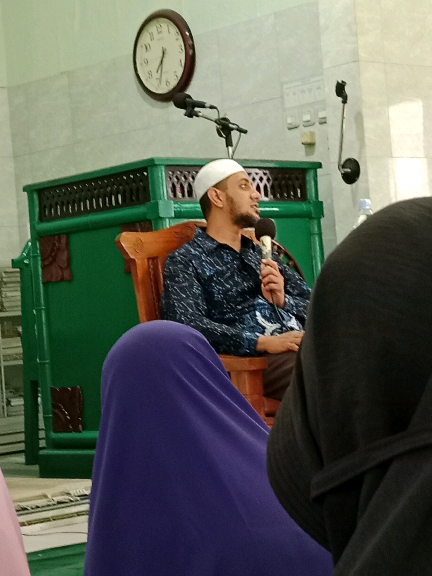 The image depicts a man wearing a white cap and a patterned shirt, seated on a wooden chair in what appears to be a room with tiled walls. He is holding a microphone and speaking, suggesting he is addressing an audience. In the background, a large green structure with intricate dark carvings is visible, and a circular clock with Roman numerals hangs on the wall. There are also two microphone stands with microphones. In the foreground, a person in a purple garment is seated, and another person in a black garment is also partially visible, suggesting a public gathering or event. The scene is likely within a mosque or community center in Cemani, Indonesia, given the context.