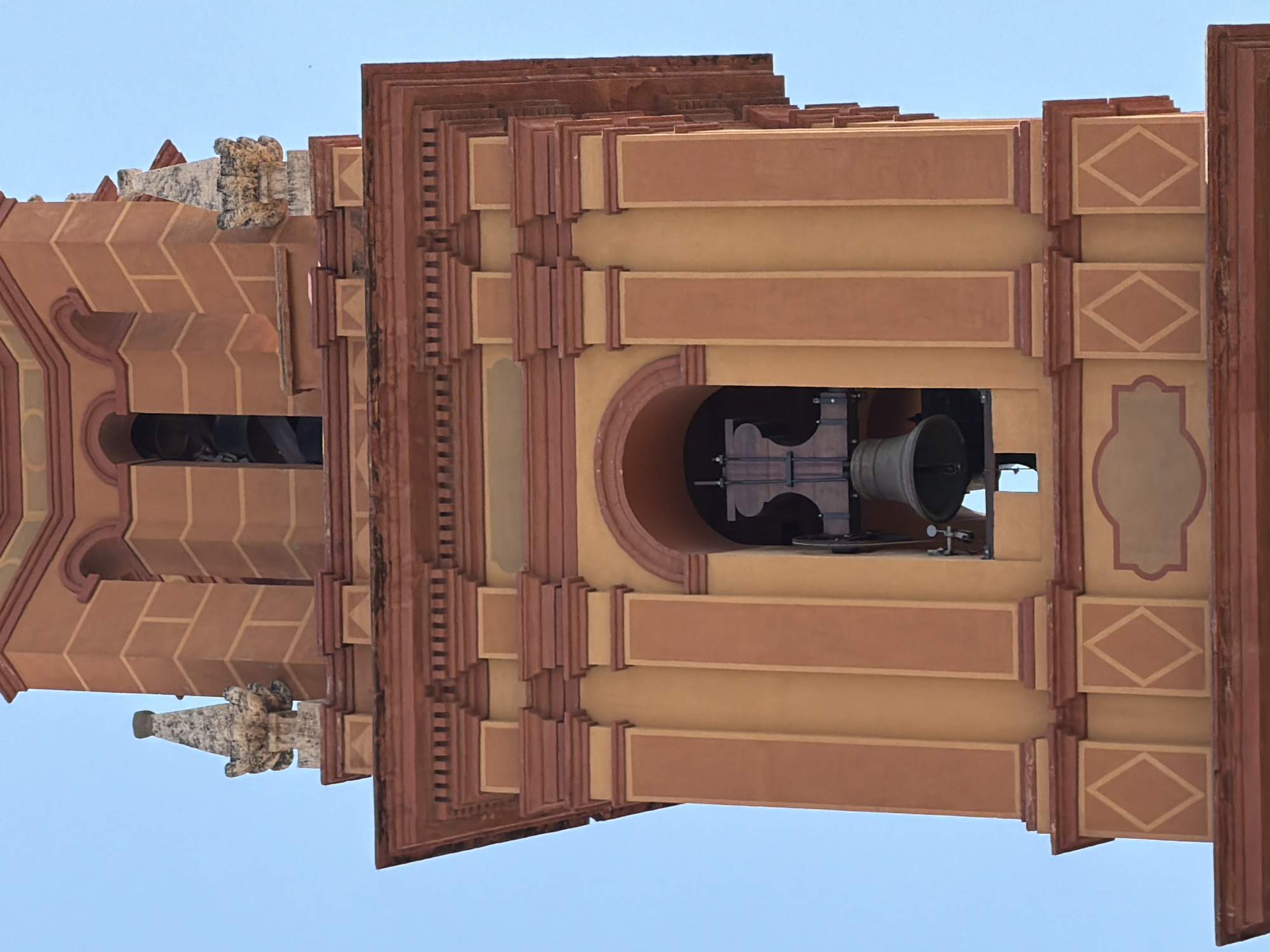 This is a close-up, upward-angle, eye-level shot of a bell tower in an unknown city. The tower is made of concrete and painted in warm tones of orange and yellow, with red accents. The architecture is ornate, featuring decorative elements like pointed finials, pilasters, and arched openings. A large bronze bell hangs within one of the arched openings, suspended from a wooden beam. The bell is ornate with horizontal bands. The sky is clear and blue.