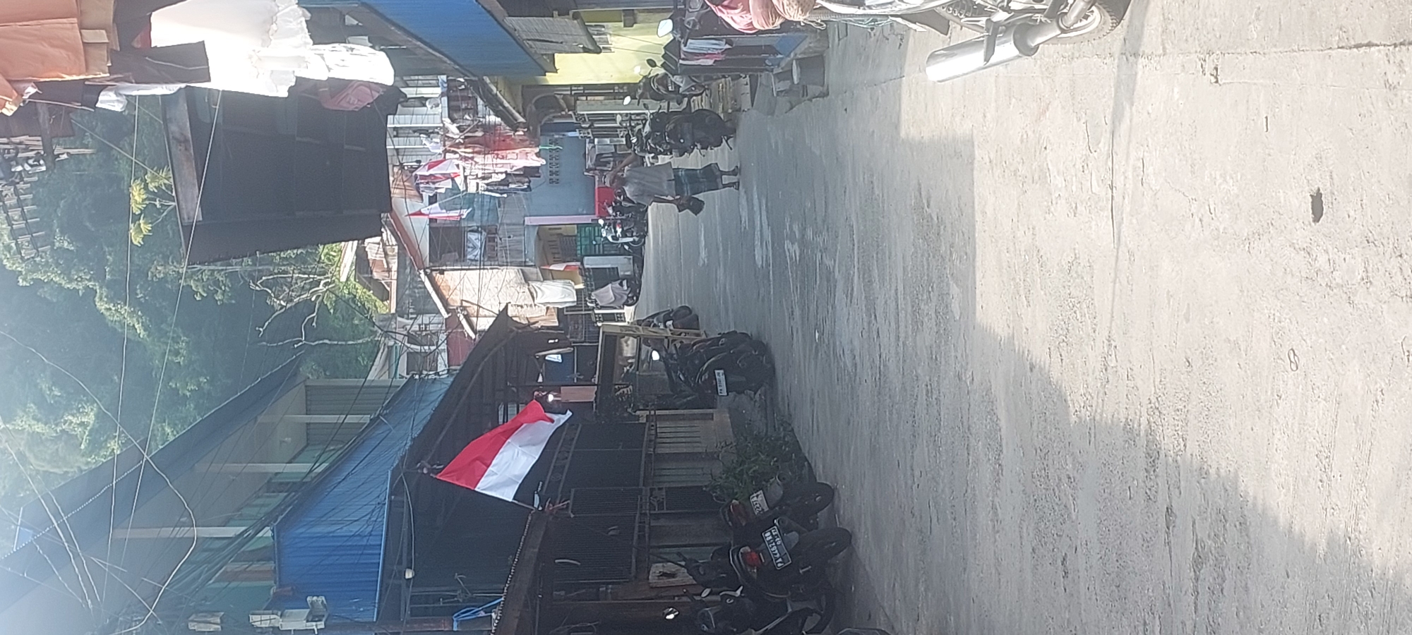 The image depicts a narrow, paved street lined with buildings on either side. The street appears to be in a residential or mixed-use area, with motorcycles parked along the sides and laundry hanging from some of the buildings. The Indonesian flag is prominently displayed on the left side of the street, suggesting a national holiday or patriotic sentiment. A man in traditional clothing is walking down the street, carrying a bag. The background shows a steep, tree-covered hillside, indicating a location with hilly terrain. Based on the presence of the Indonesian flag and the visual characteristics of the buildings and landscape, the scene is likely located in Jayapura, Indonesia, a city known for its coastal location and hilly topography.