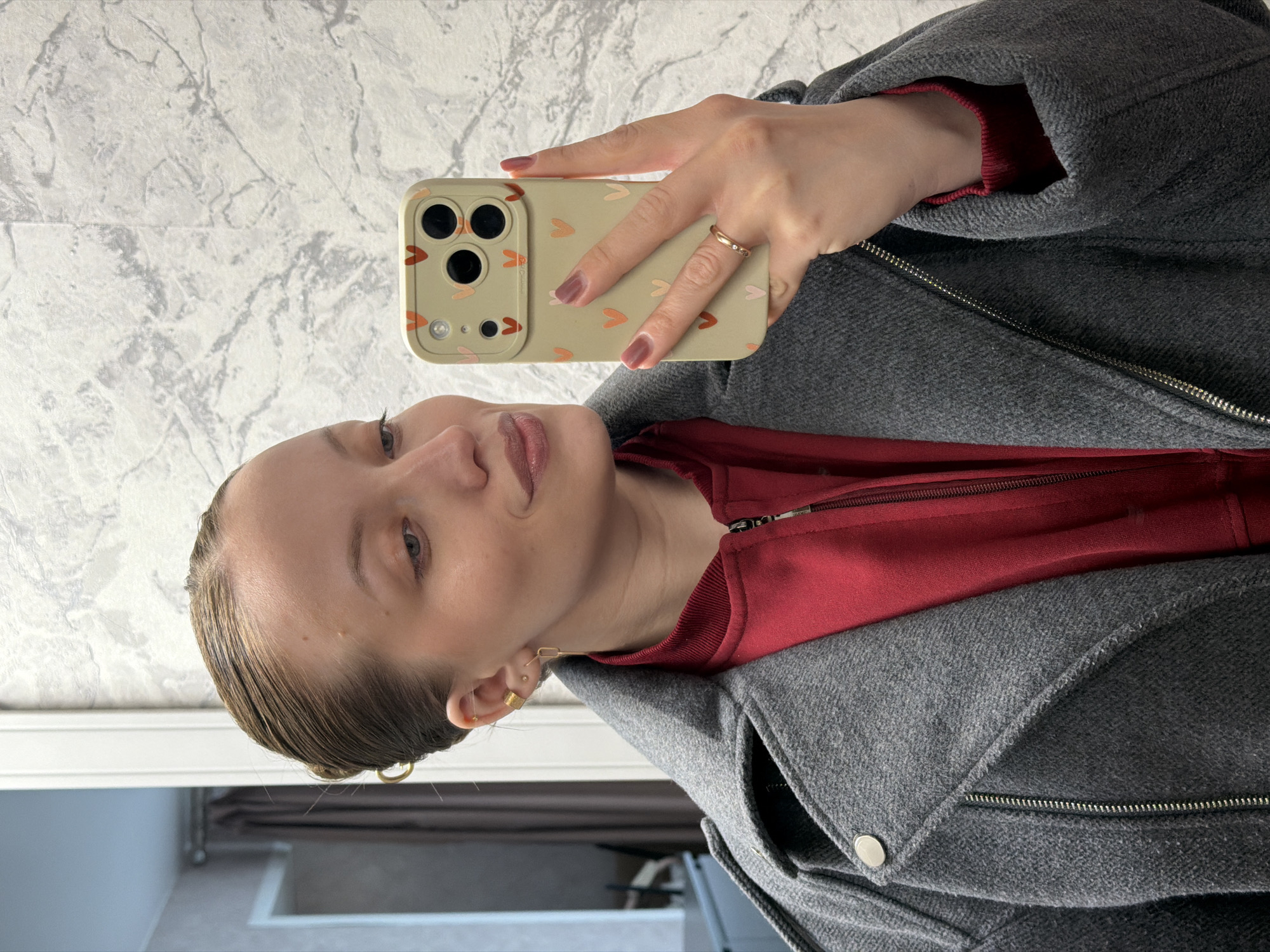 A woman takes a selfie with a beige smartphone adorned with a heart pattern on its case. She is wearing a red zip-up jacket and a gray wool coat. She has fair skin, light brown hair pulled back, and is wearing small gold earrings and a gold ring on her ring finger. The background appears to be an interior wall with a marble-like pattern. The location is unidentifiable as Reshetnikovo, Russia.