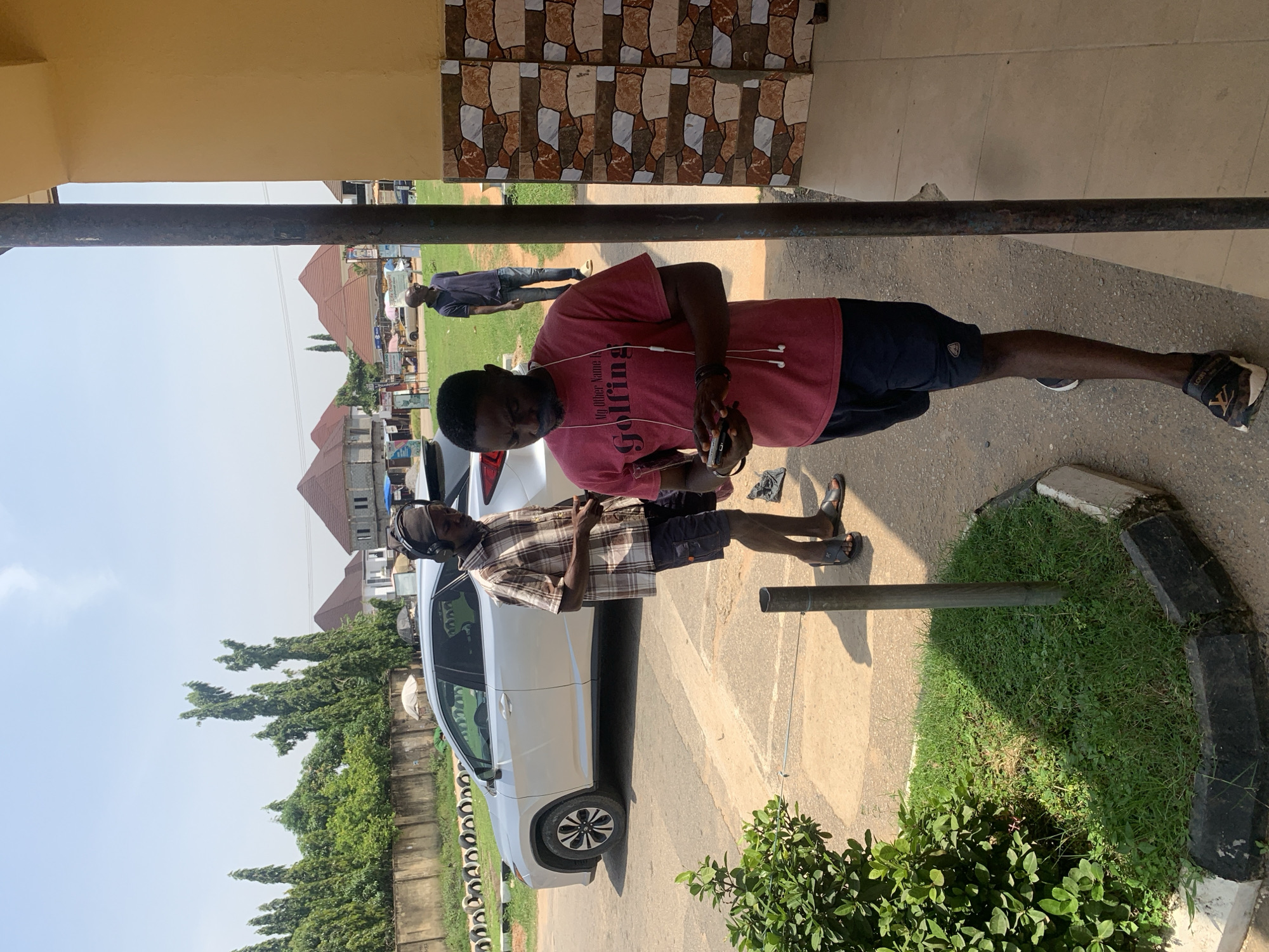 In Abuja, Nigeria, on a sunny day, a light-colored SUV is parked on the side of a paved road. Two men are visible. One man, wearing a plaid shirt and dark shorts, stands near the SUV, holding headphones over his ears and looking down. The other man, in a red t-shirt and dark shorts, stands closer to the foreground, holding a phone in his hands and appears to be using it. Behind them, several houses with red roofs are under construction, and lush green trees line the background. A stone wall with a decorative tire border runs along the left side of the road. The scene is set in an urban area with signs of ongoing development.