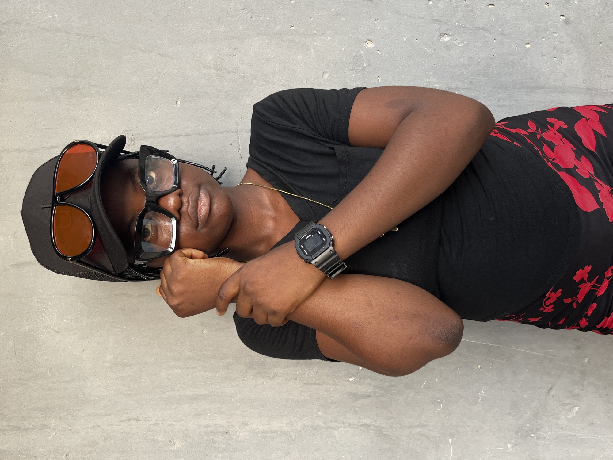 An individual, wearing a black t-shirt and a black skirt with red floral prints, is lying on a light gray surface. The person has on a black baseball cap, with sunglasses on the brim, and glasses over their eyes. A digital watch is on the left wrist. The location is in Badagry, Nigeria, but the precise location within the city is indiscernible.