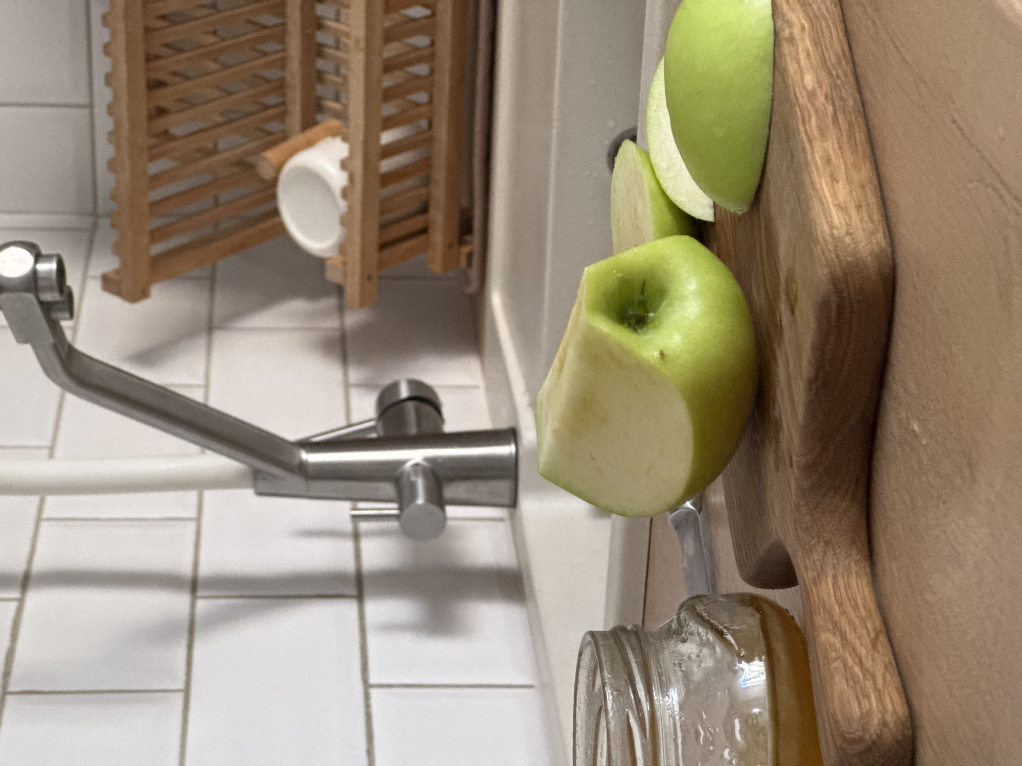 This is a close-up, low-angle shot of a kitchen counter in Mirnoye, Ukraine. In the foreground, a wooden cutting board holds a partially sliced green apple and two apple slices. To the left of the cutting board, a glass jar filled with honey sits on the counter. In the background, a stainless steel faucet extends over a white sink, with a wooden dish rack holding a white bowl and other items visible behind it. The wall behind the sink is tiled with white subway tiles. The overall scene suggests food preparation is underway or has recently occurred. There are no people present in the image.