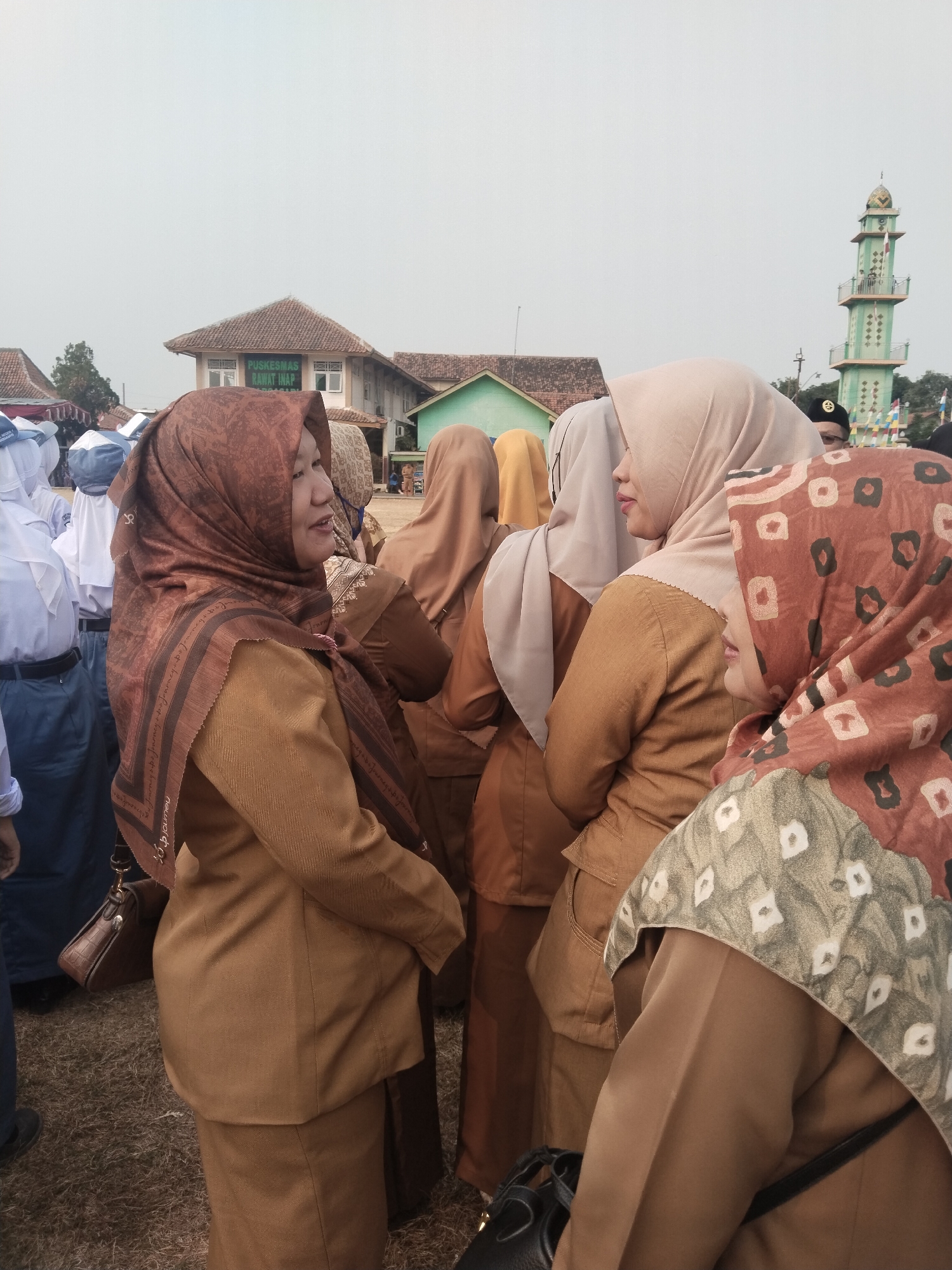 The image captures an outdoor gathering in what appears to be a village in Pakulaut, Indonesia. In the foreground, four women in light brown uniforms and hijabs are engaged in conversation. One woman, on the left, wears a patterned brown hijab and a brown jacket, and has a small brown handbag hanging from her left shoulder. To her right, another woman in a beige hijab and brown jacket is speaking. Further to the right, a third woman in a beige hijab and brown jacket looks away, while a fourth woman, partially obscured, wears a patterned brown hijab with a geometric design. Behind them, a group of individuals in white uniforms and blue head coverings are visible, possibly students. In the background, a building with a sign that reads "PUSKESMAS RAWAT INAP" (a type of clinic) is seen, along with a green mosque minaret. The ground is covered with dry grass and dirt. The overall atmosphere suggests a formal event or gathering.