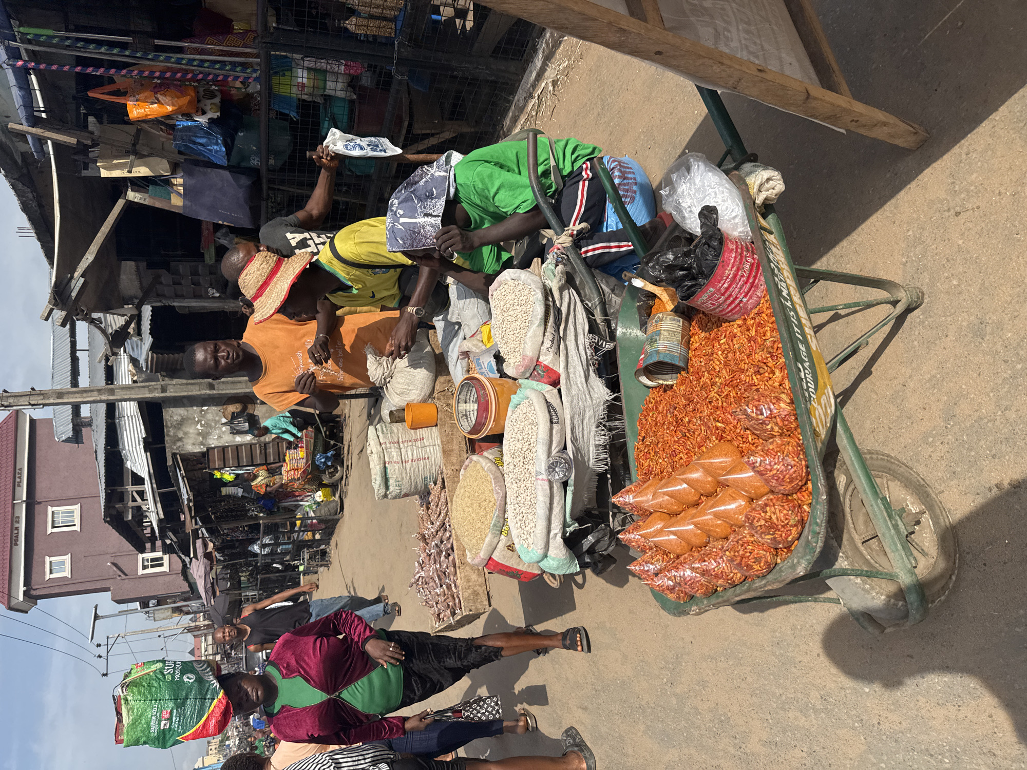In an outdoor market in Onitsha, Nigeria, a street vendor and customers interact at a handcart. The handcart is filled with spices, dried goods, and bags of red peppers. Three men are present at the cart: one, wearing an orange shirt and straw hat, appears to be the vendor. Two other men are crouched over the cart, examining the goods. Various bags and sacks of ingredients are on display. In the background, other people and buildings are visible. A woman passes by carrying a green bag on her head. The setting shows a typical African market scene.