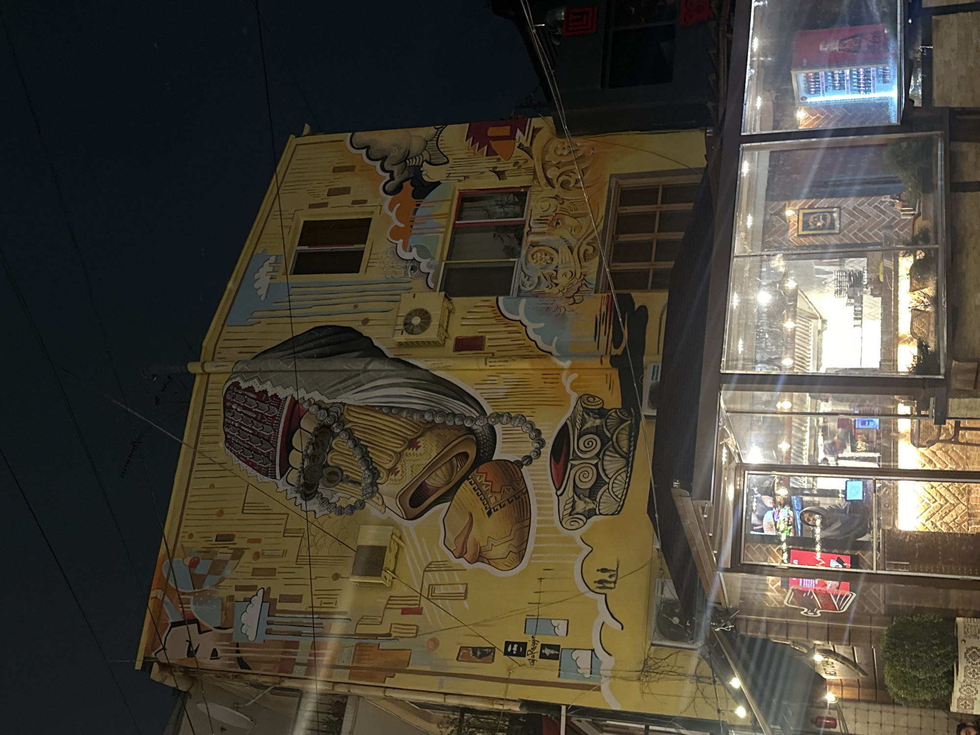 The image shows a building in Yerevan, Armenia, featuring a large mural painted on its side. The mural is predominantly yellow and depicts a series of stylized objects or figures, including what appears to be traditional Armenian items or symbols. The building is adjacent to a storefront with large glass windows, reflecting the interior. A person is visible inside the store. The sky is dark, suggesting it is either night or dusk. Overhead power lines are visible in the sky.